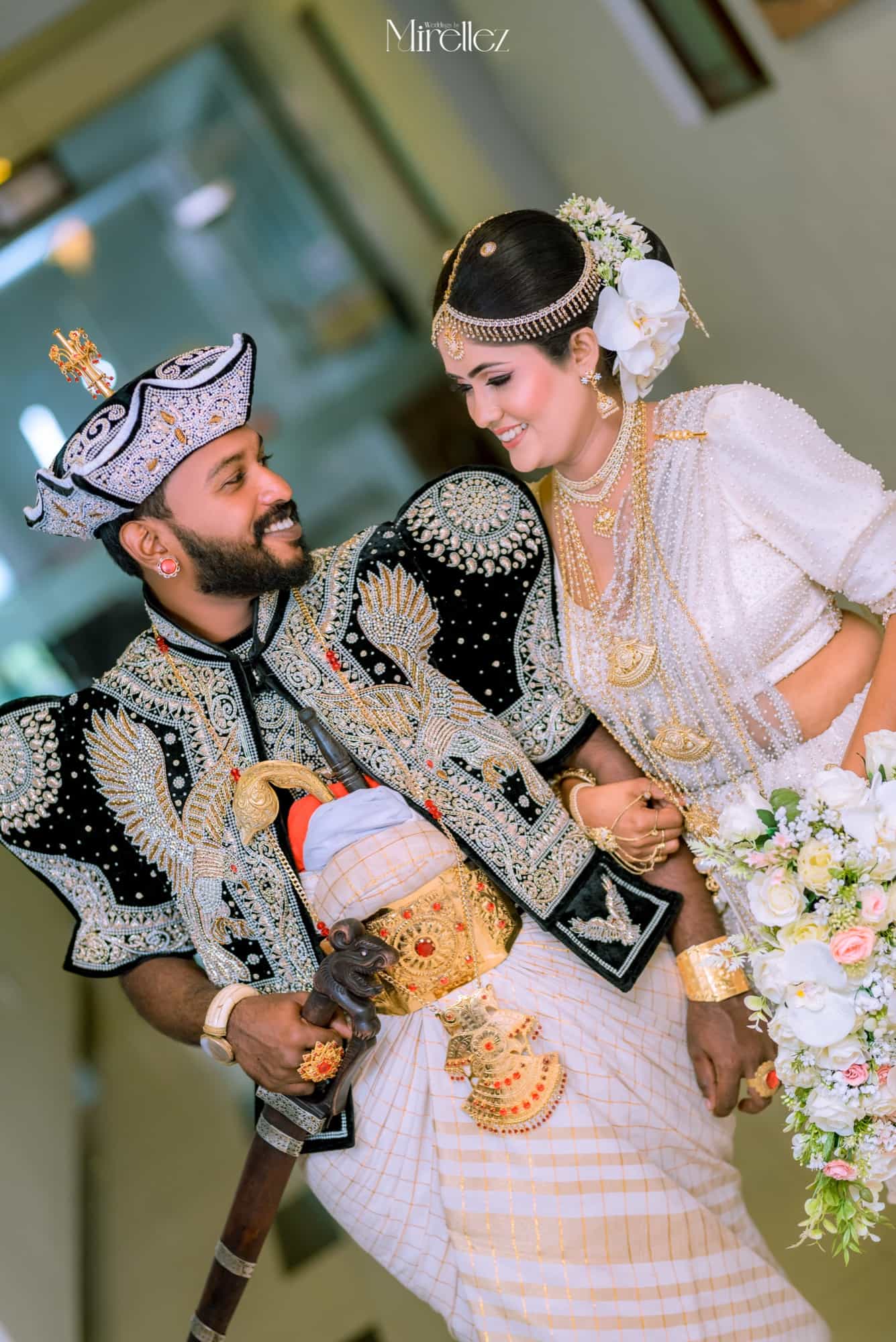 The groom in full Kandyan nilame dress turns to smile at his bride as she laughs, her cascading white rose and orchid bouquet at her side