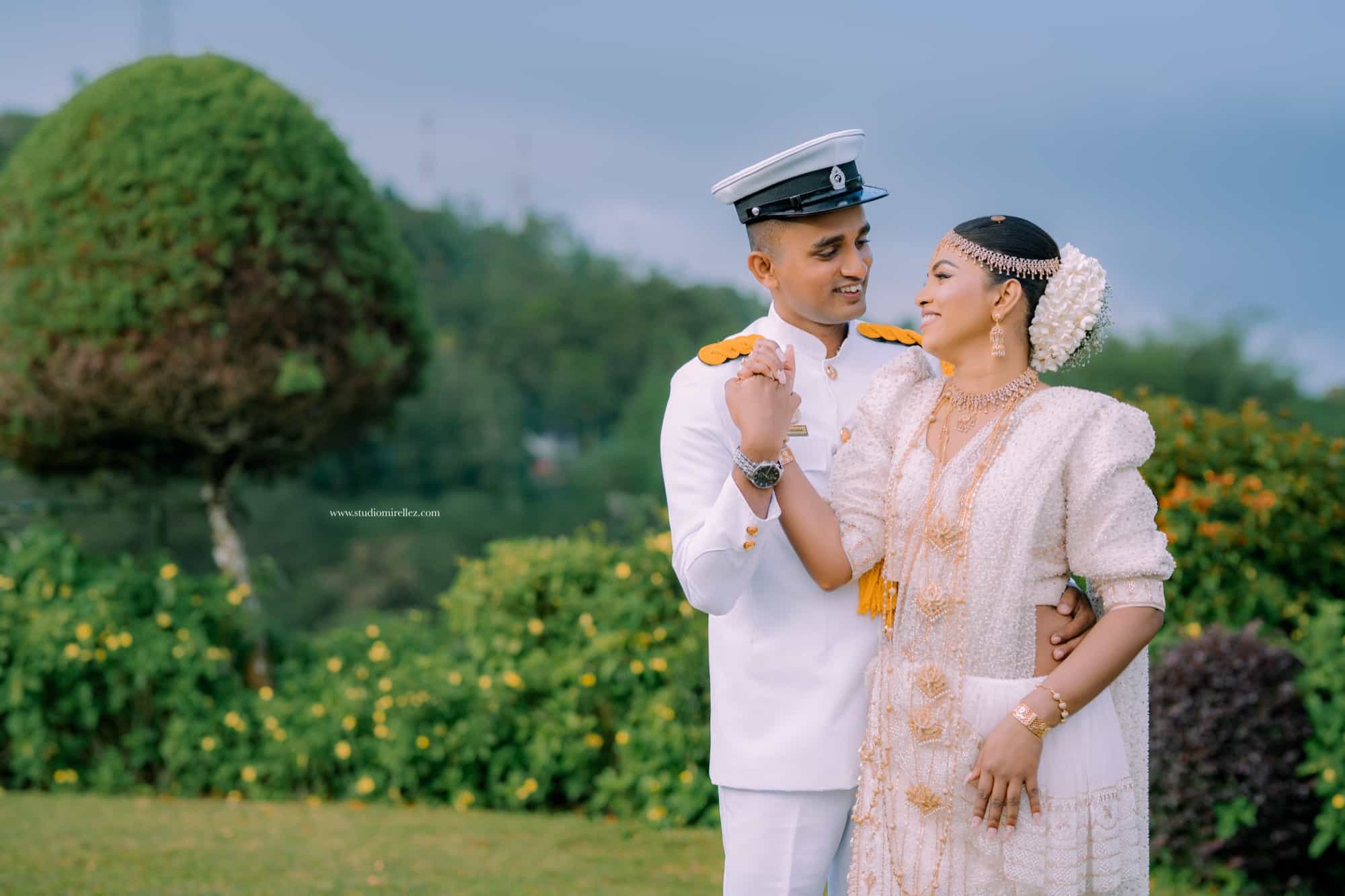 Groom in naval whites raises the bride's hand as if to twirl her, both smiling beside a topiary tree under an overcast sky