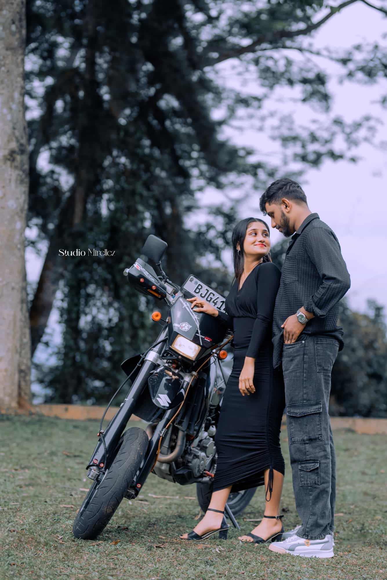 Couple in matching black outfits beside a motorbike on a misty hillside lawn, she leans back against him smiling