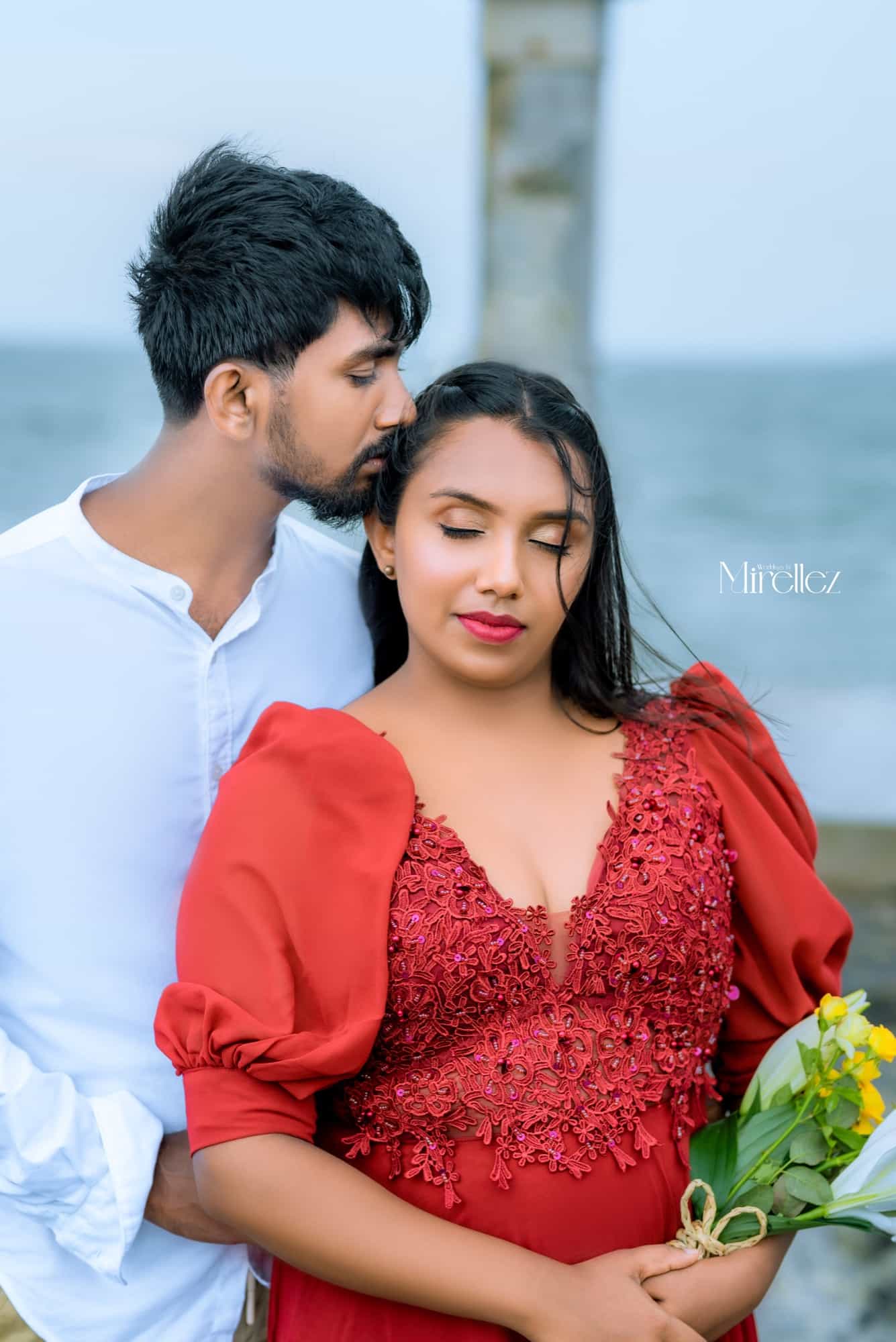Akash kisses Hansi's temple as she closes her eyes, holding a bouquet of yellow blooms, the sea soft behind them