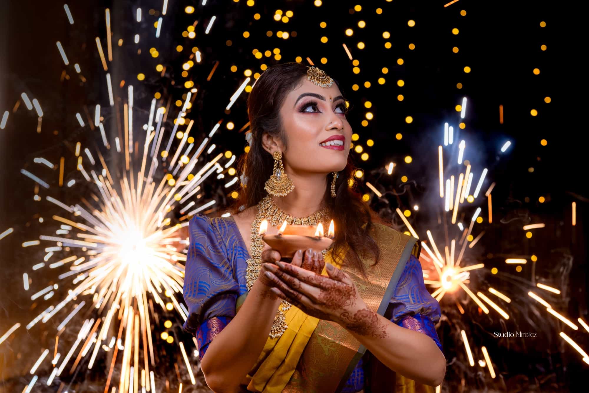 Woman in a yellow and blue Kanjivaram saree cradling a lit clay diya, framed by bursting sparklers and fairy lights