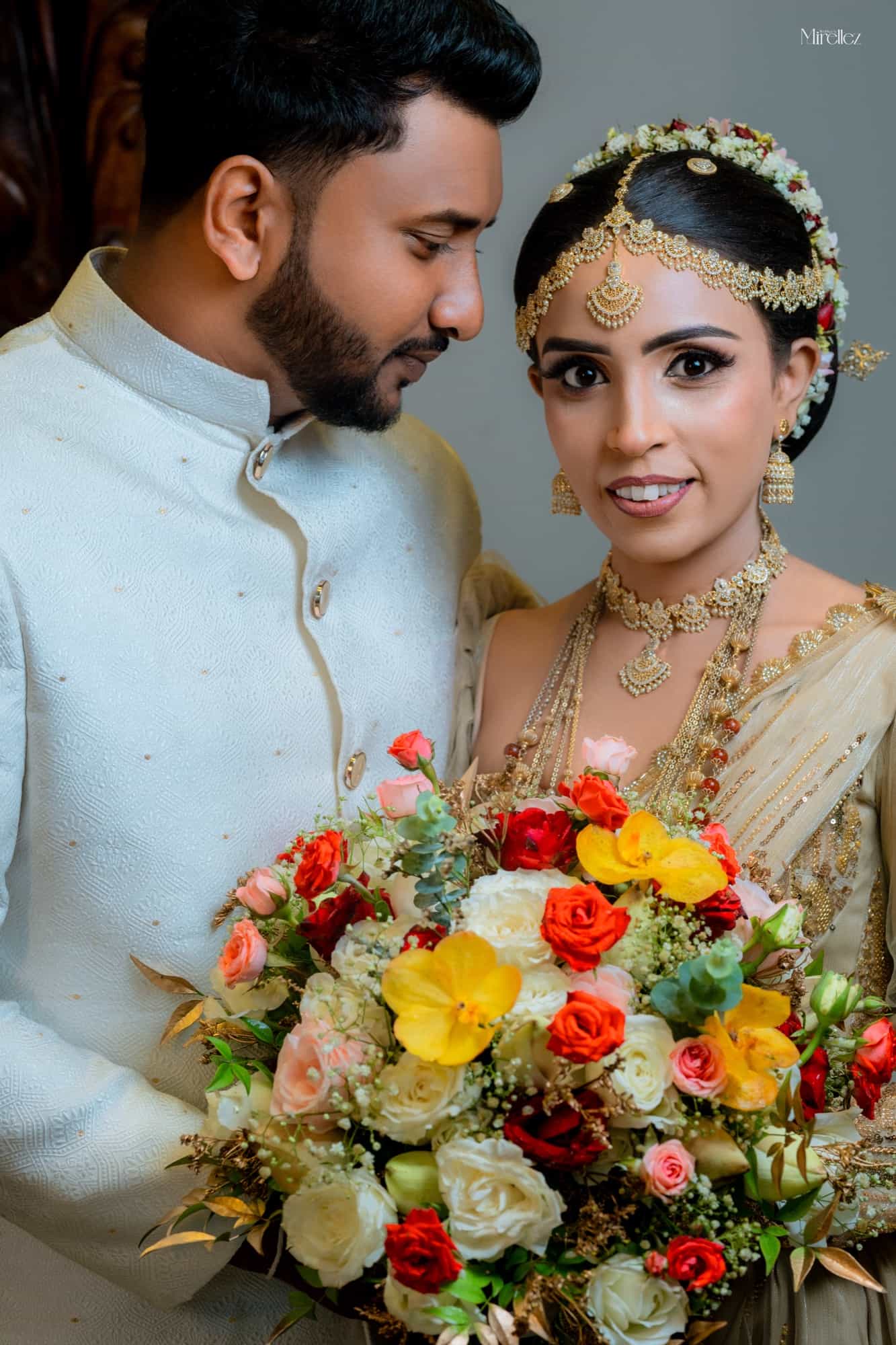 Bride in a gold-embroidered Kandyan saree looks to camera while the groom in a cream sherwani gazes at her, a rose bouquet between them
