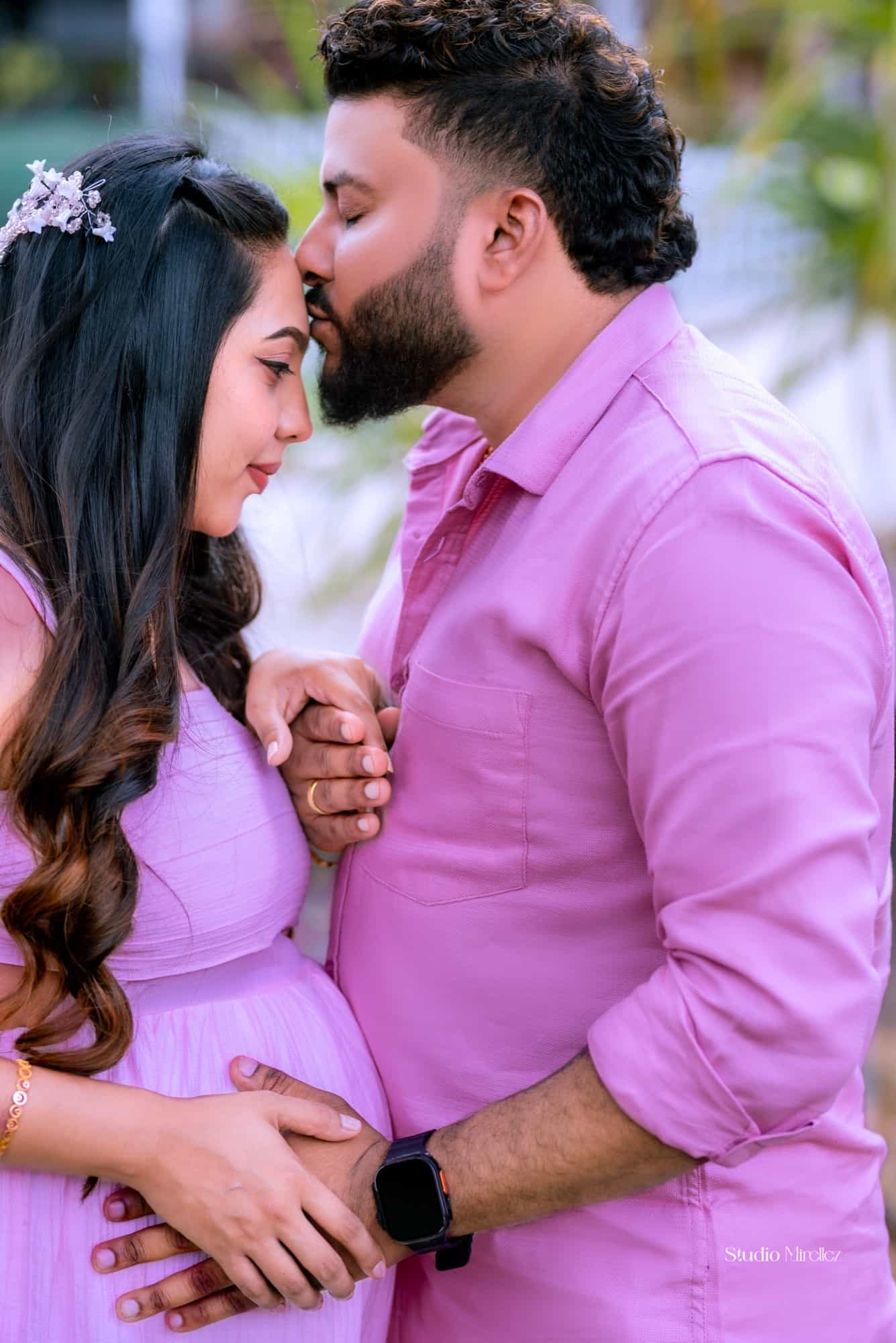 Expectant mother in a lilac dress leans into her husband as he kisses her forehead, hands resting on her bump