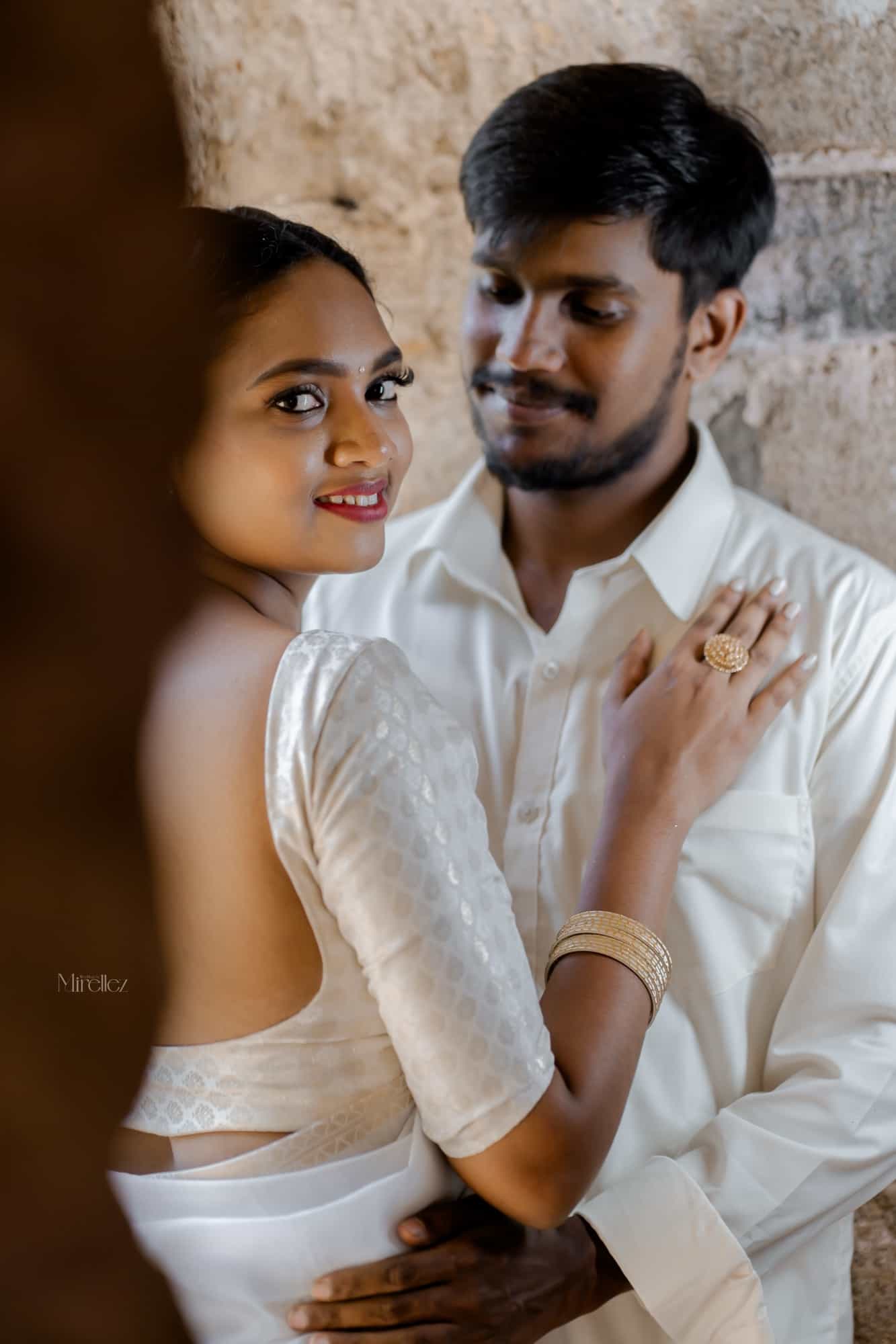 Bride in a white silk saree glances back over her shoulder as her groom holds her from behind against a pale stone wall