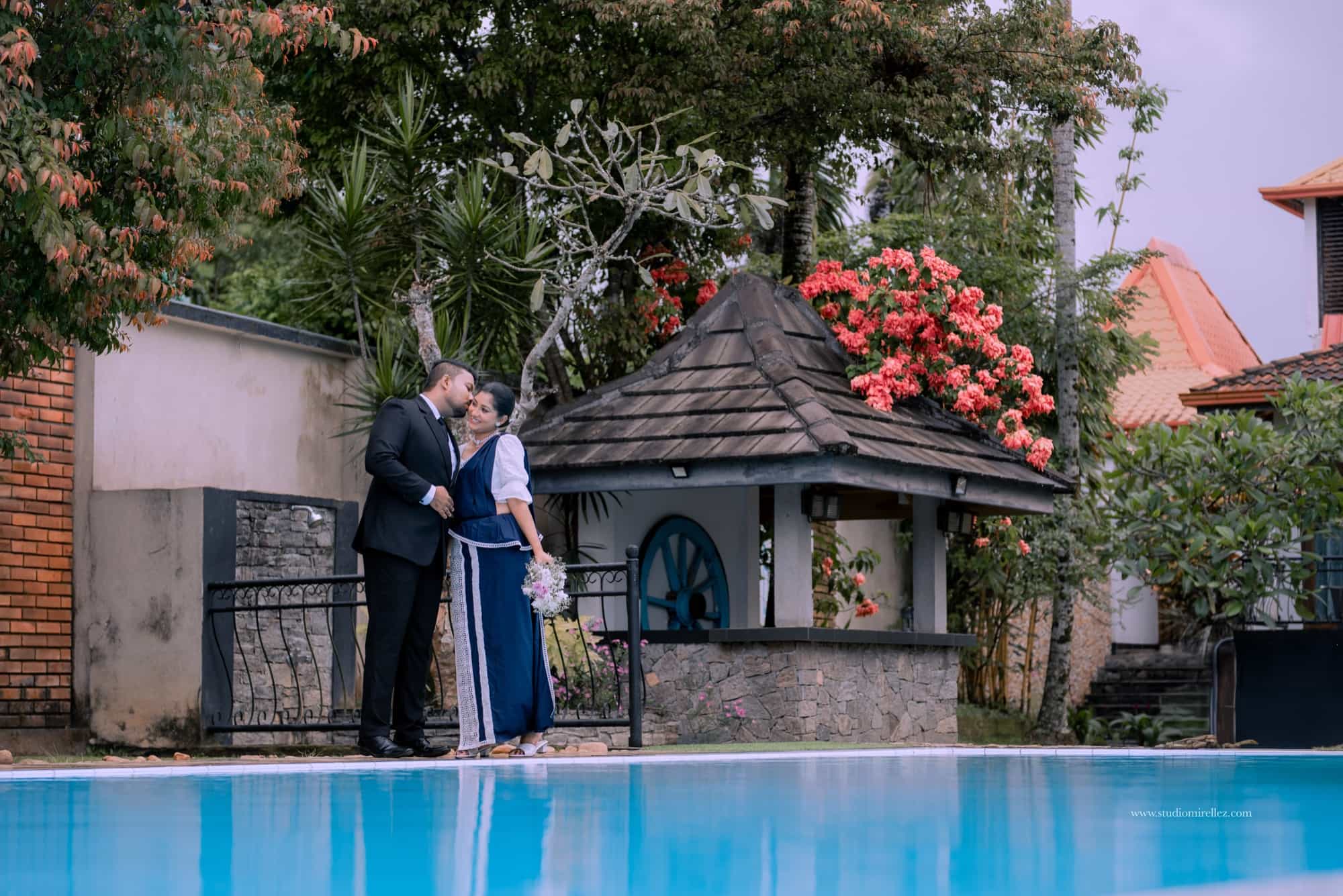 Wide poolside frame of the groom kissing his fiancee's forehead beside a tiled cabana, red bougainvillea and turquoise water