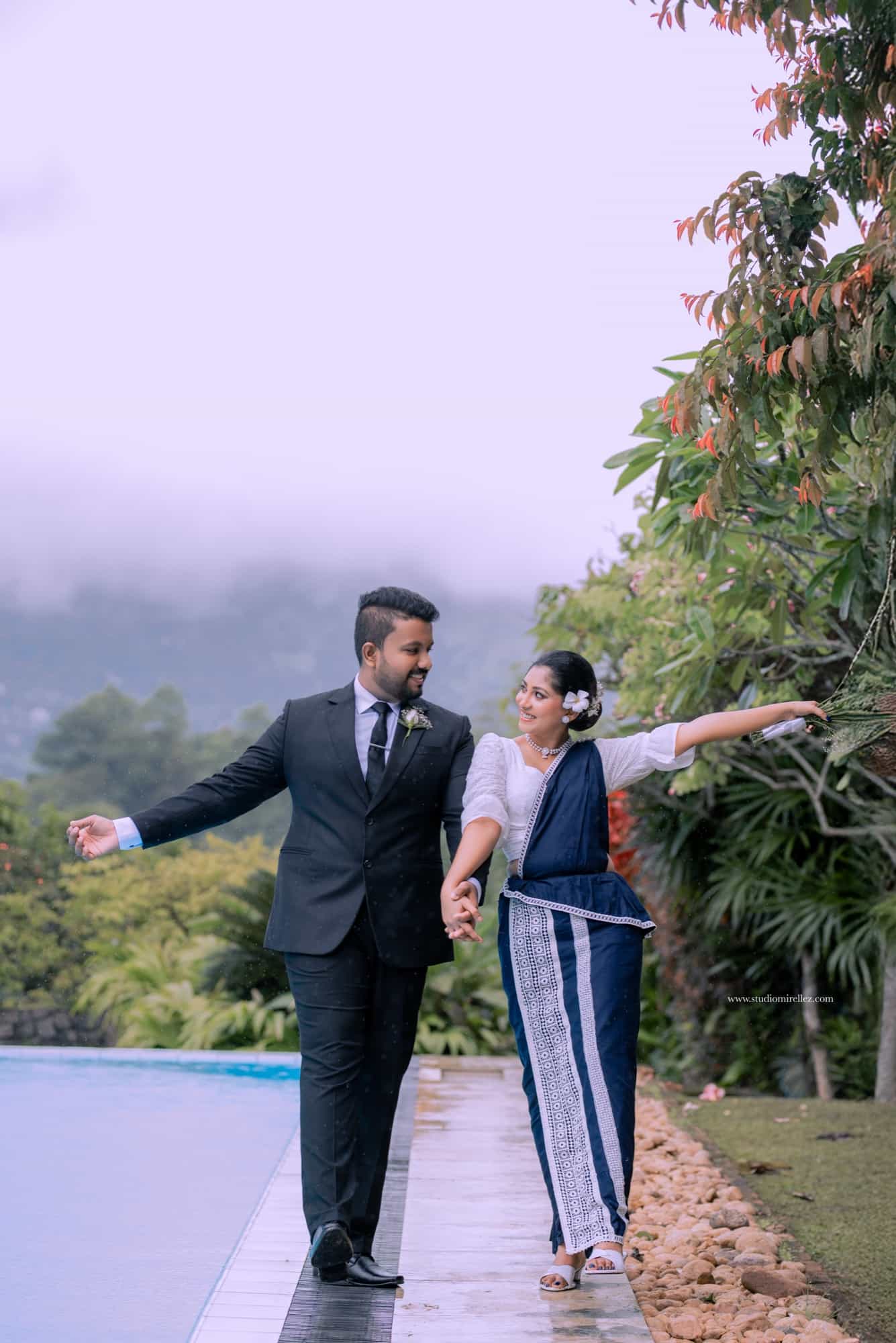 Couple walking hand in hand along a wet infinity-pool edge in light rain, mist-covered hills behind them