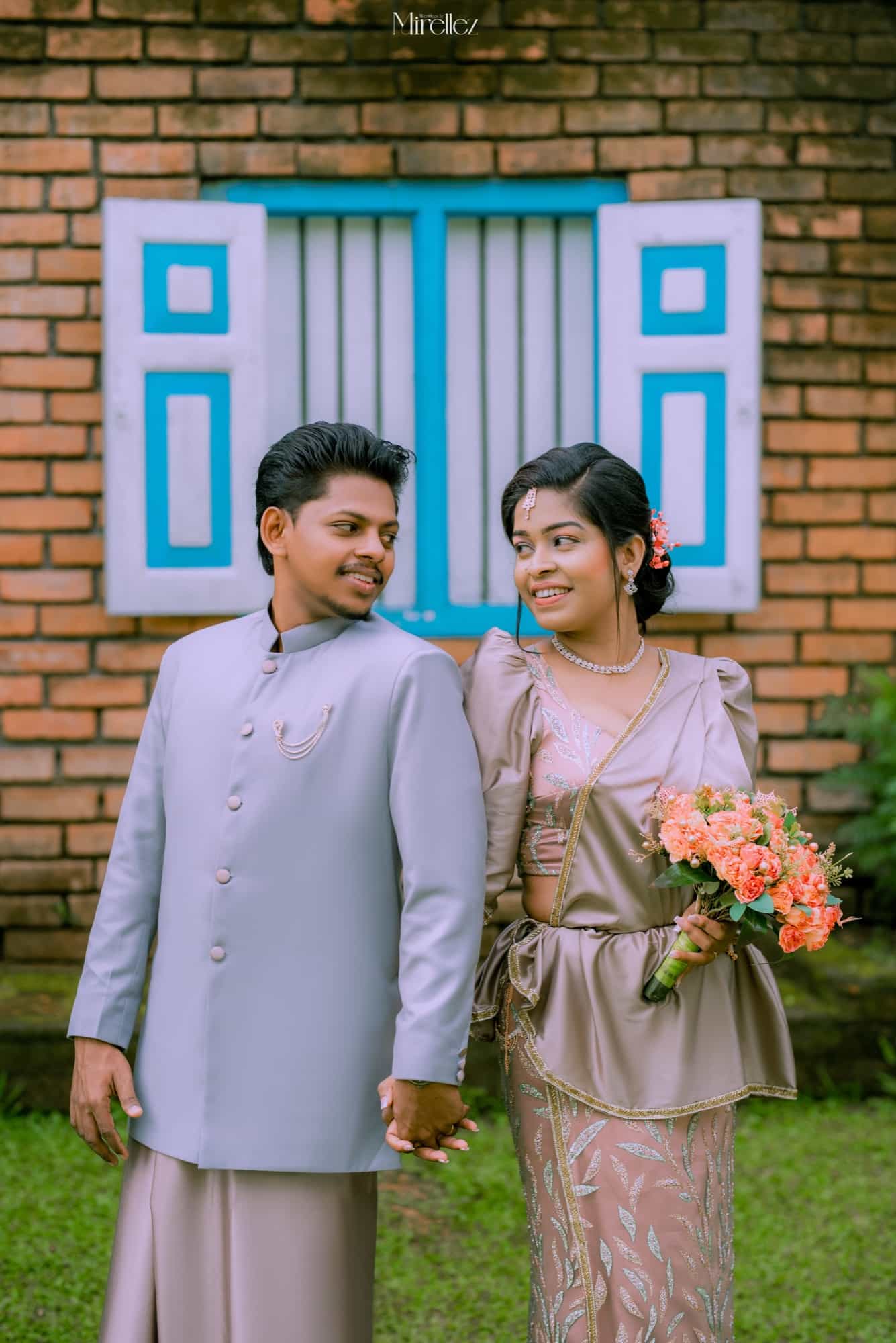 Karthik and Anjani hold hands before a red brick wall with a bright turquoise shuttered window, she smiles up holding a coral rose bouquet