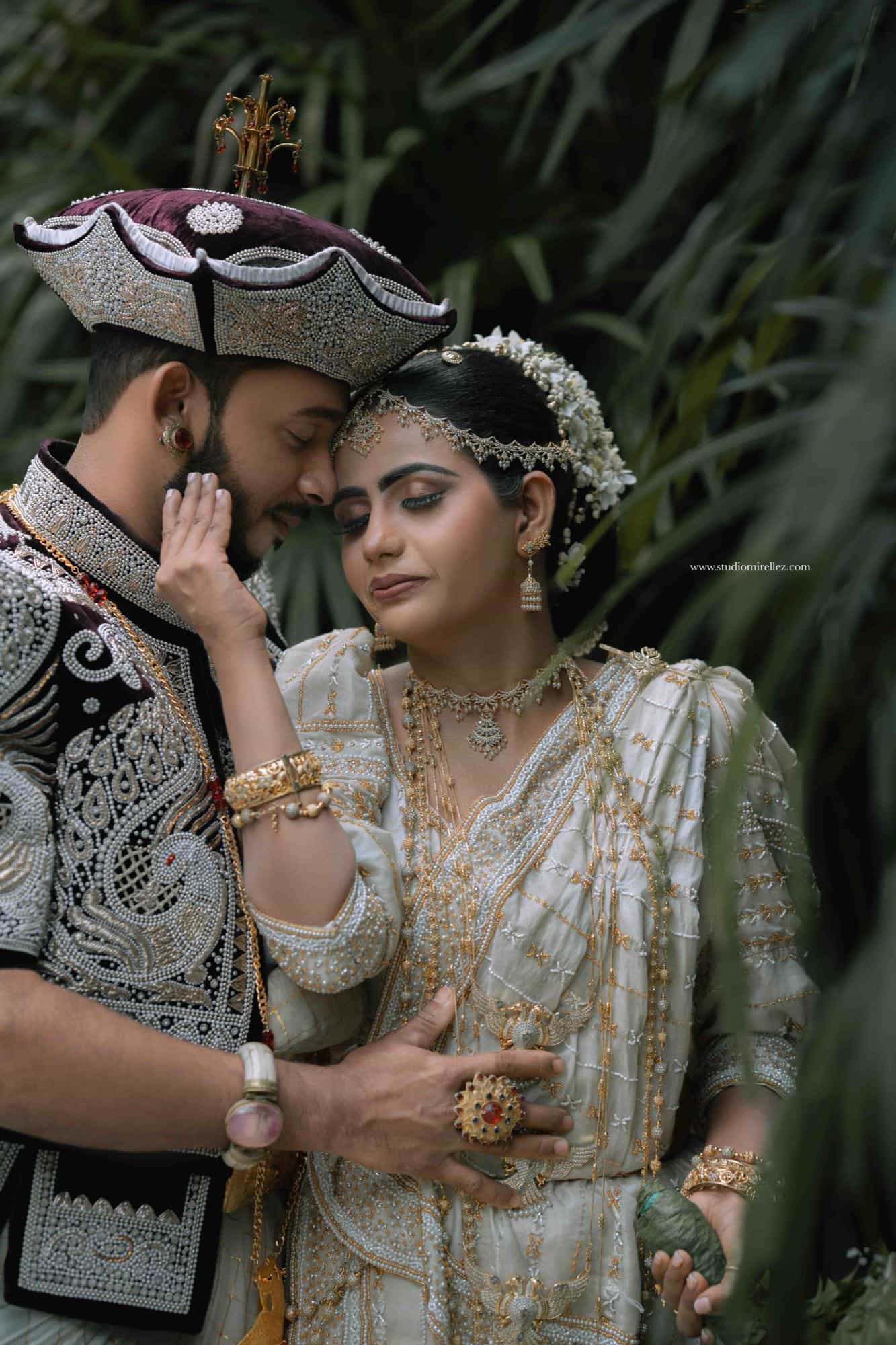 Bride and groom forehead to forehead among dark garden foliage, he in a jewelled nilame headdress, she in a white and gold osari with a pearl headpiece