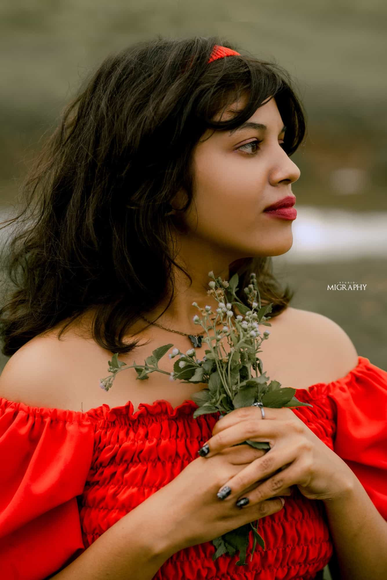 Close-up profile portrait of Kawya in a red off-shoulder top with wind-swept hair, holding small white wildflowers