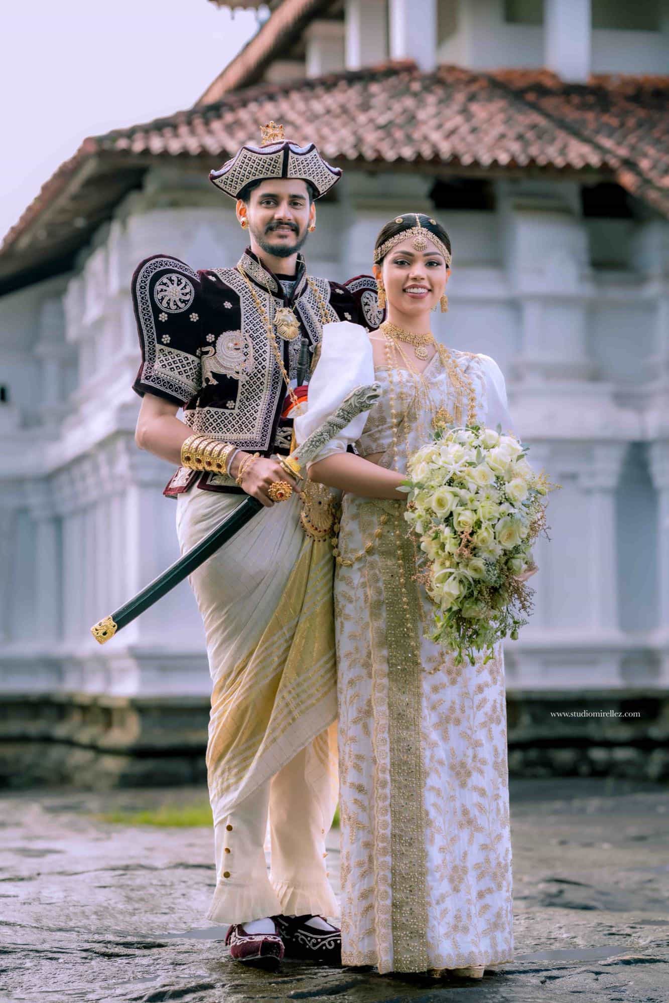 Full-length portrait of the couple before a white and terracotta temple, the groom carrying a ceremonial sword, the bride holding a cascading white rose bouquet