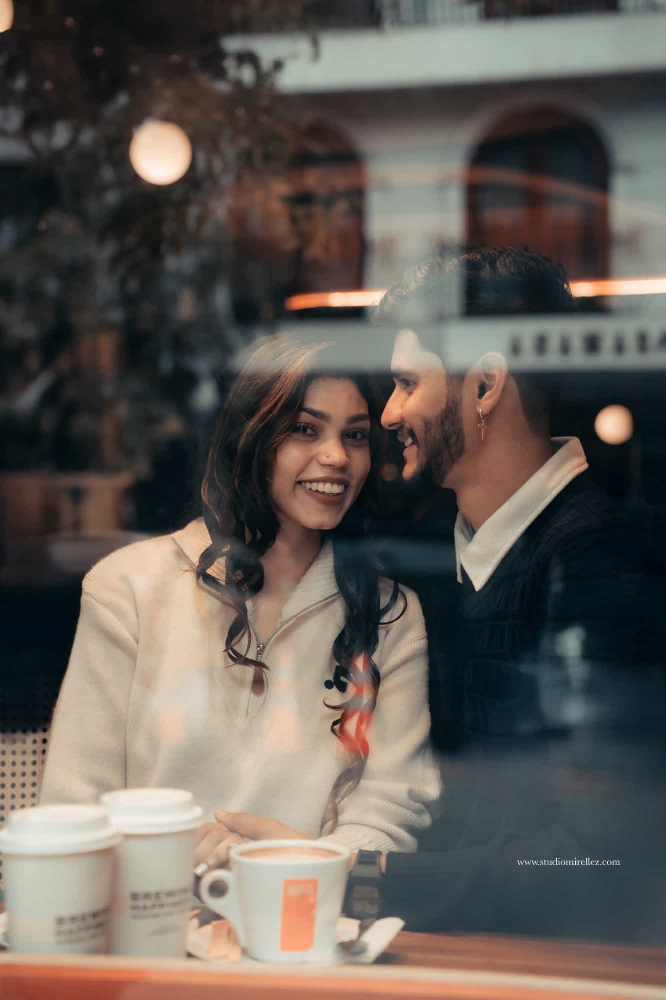 Couple seated inside a cafe photographed through the window glass, city reflections layered over them as she laughs