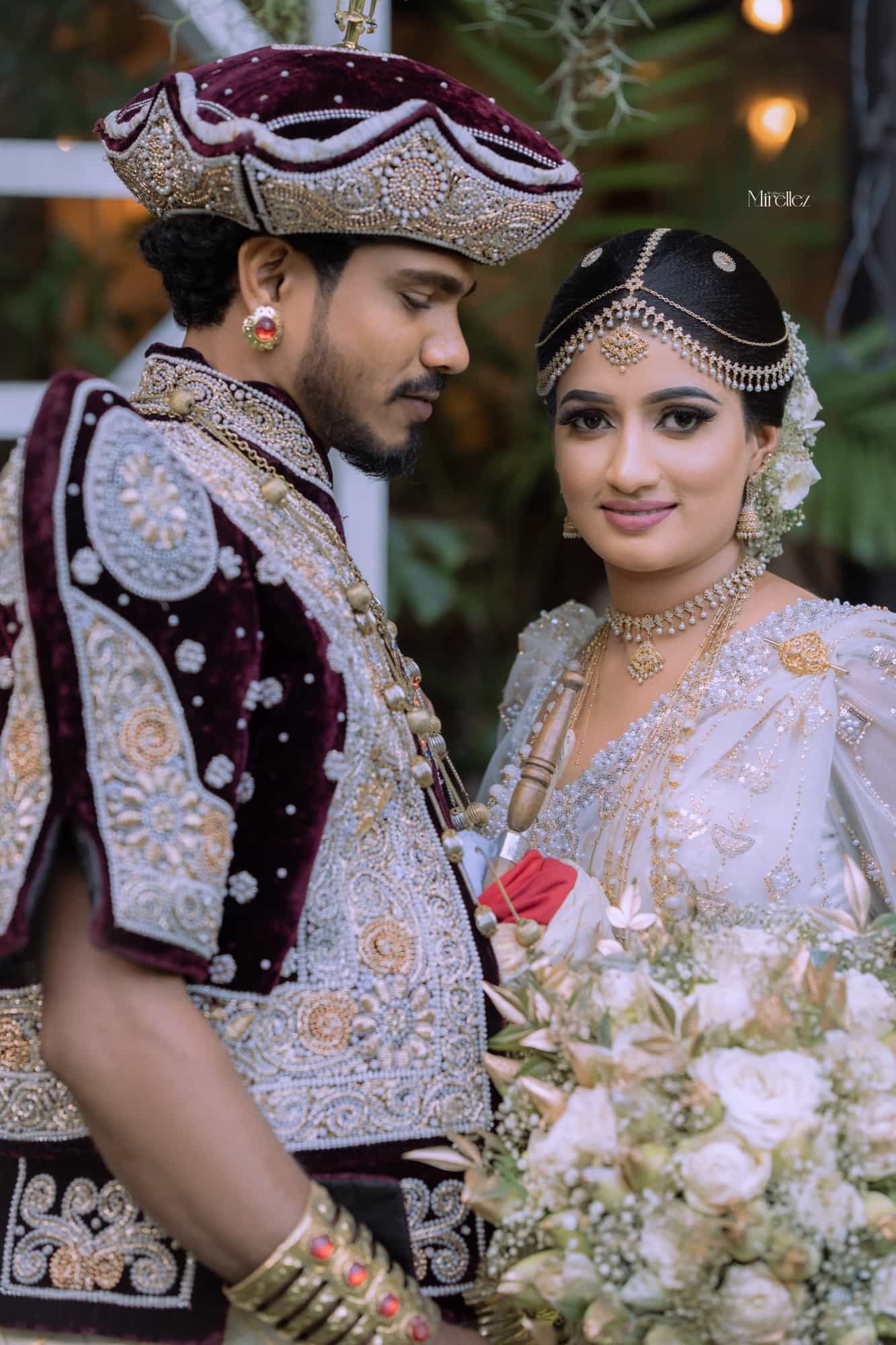 Close portrait of the bride in a beaded white Kandyan saree and pearl headpiece, the groom in a maroon velvet nilame jacket turned toward her