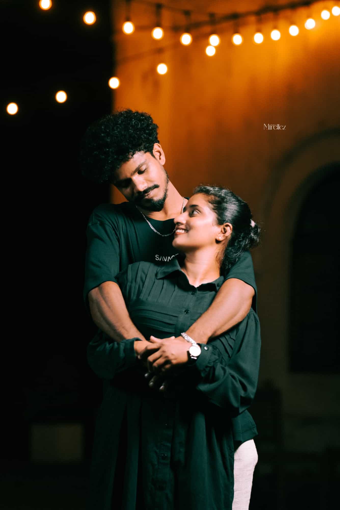 Couple in matching dark green embrace beneath warm bistro string lights, she looks up at him against a glowing amber wall