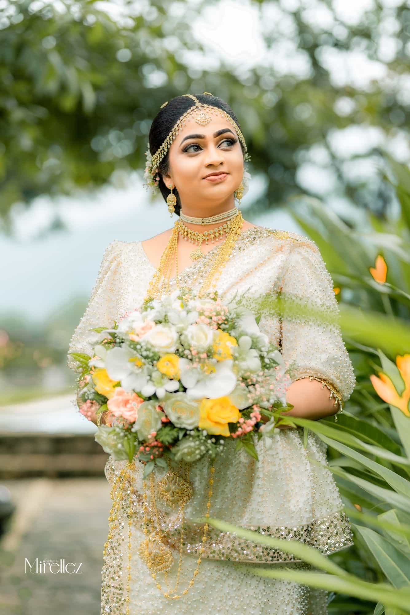 The bride alone in a pearl-beaded white osariya and gold headpiece, holding a bouquet of white and yellow roses against green foliage