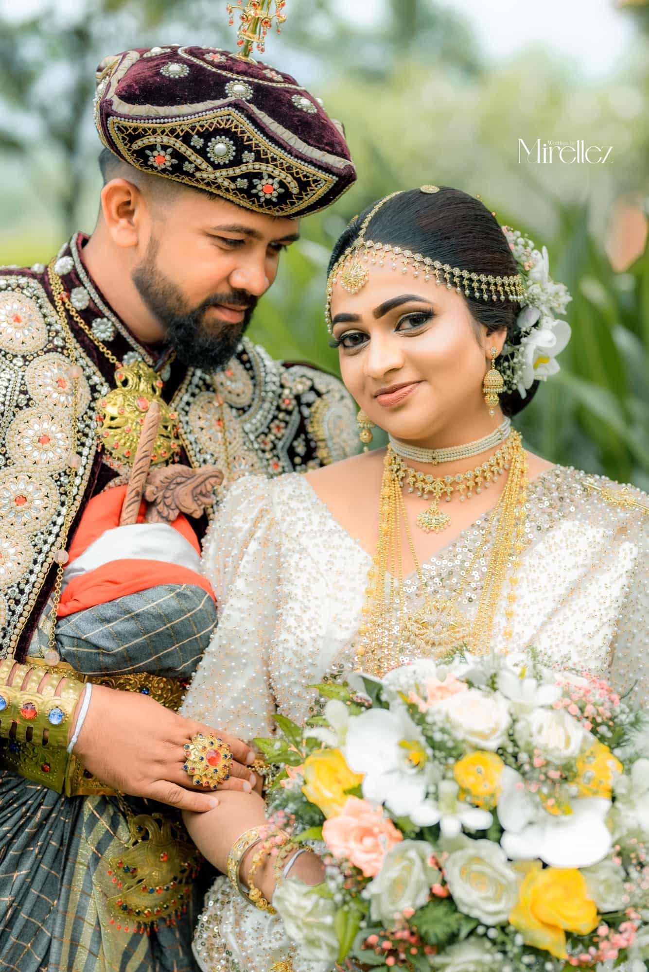 Close portrait of the bride and groom in Kandyan wedding dress, the groom in an embroidered velvet headdress, the bride looking softly to camera
