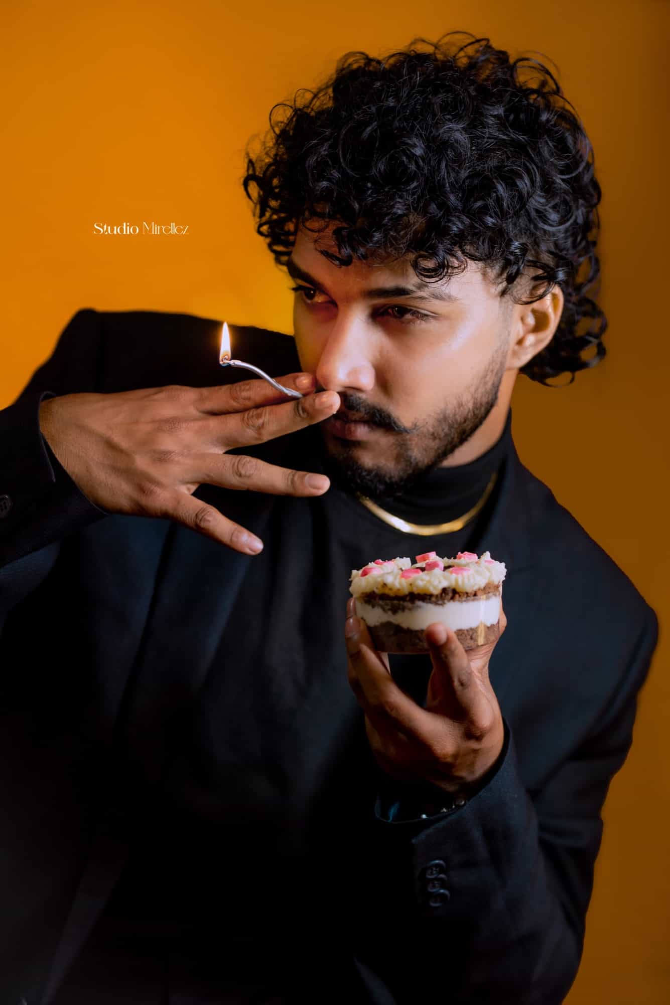 Poorna in a black suit lifts a lit birthday candle away from a small layered cake, glowing against a warm amber backdrop