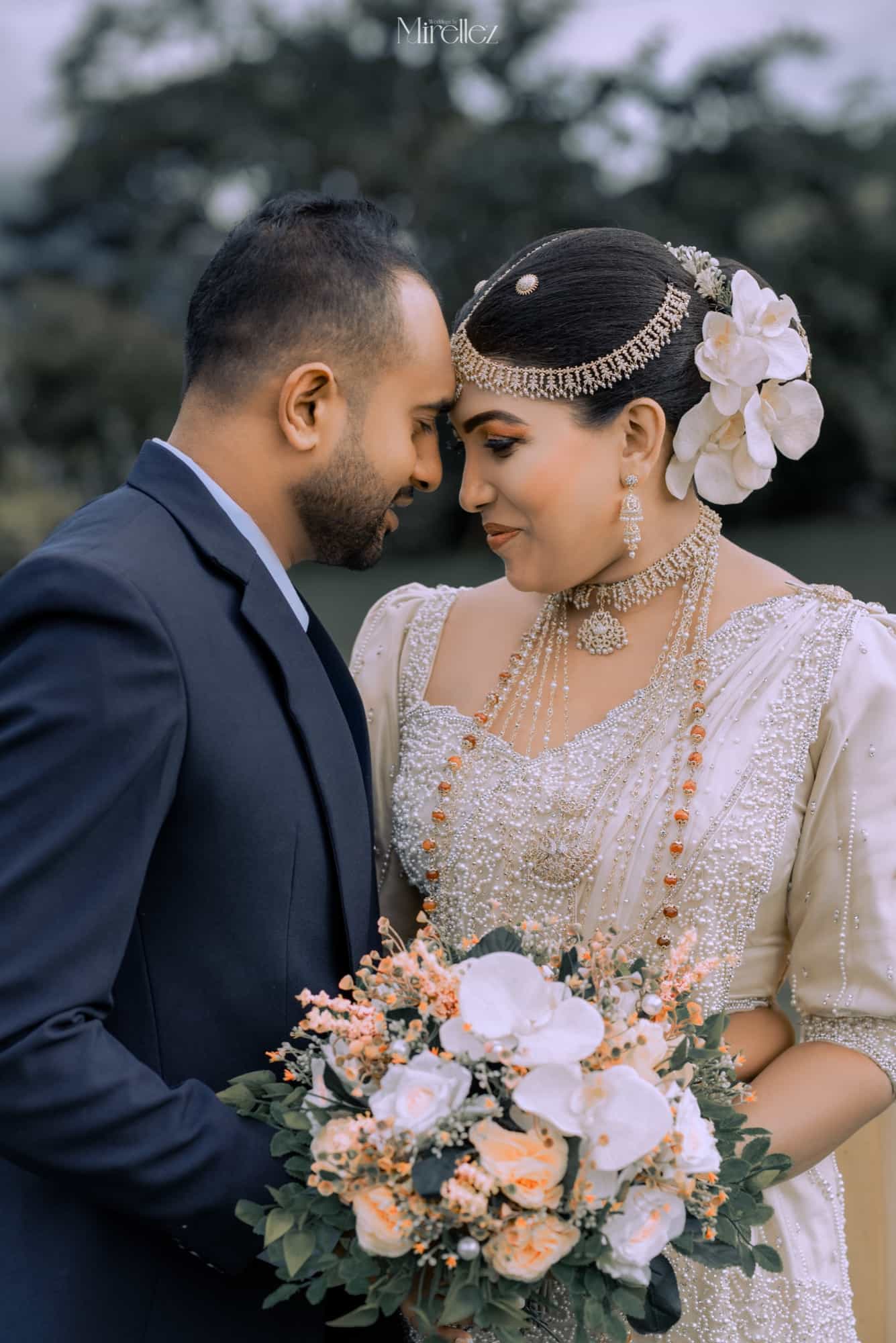 Close profile of the couple touching foreheads, the bride wearing a pearl headpiece and white orchid, a peach bouquet between them