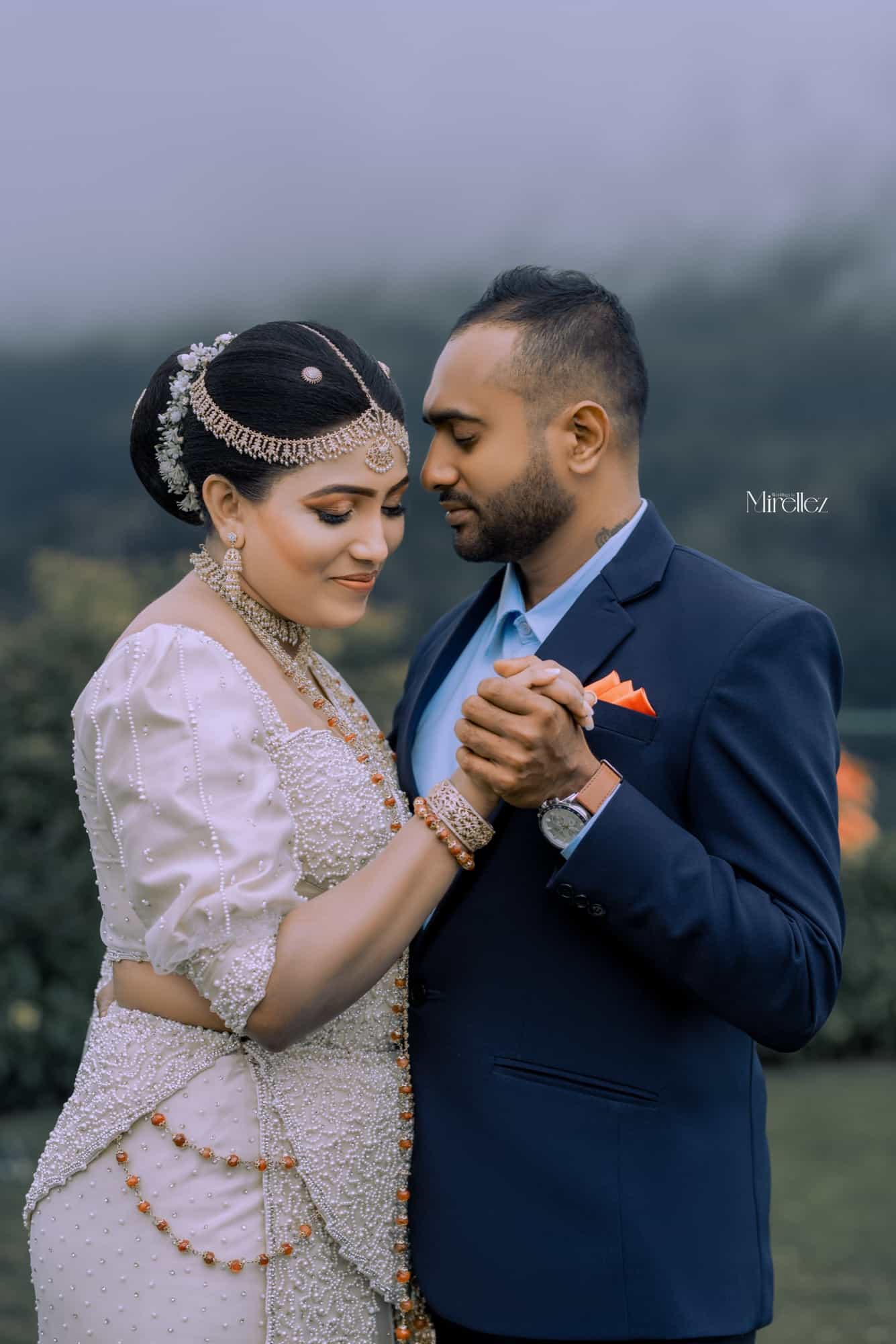 Bride in a pearl-beaded Kandyan osariya and groom in a navy suit stand close, hands clasped, against a misty hillside