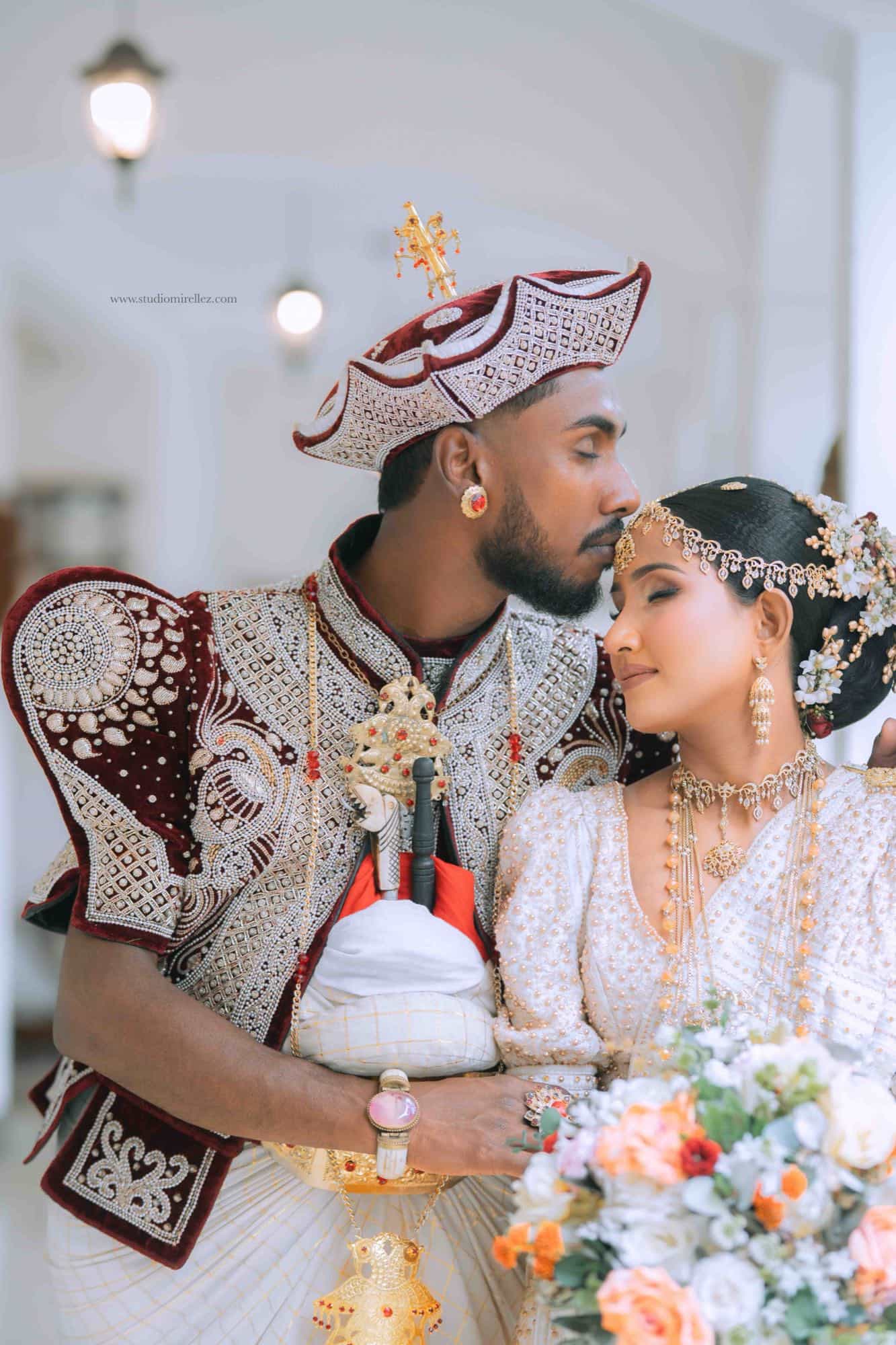 The groom in a maroon and white Kandyan jacket kissing the bride's forehead, her peach and cream bouquet raised in the foreground