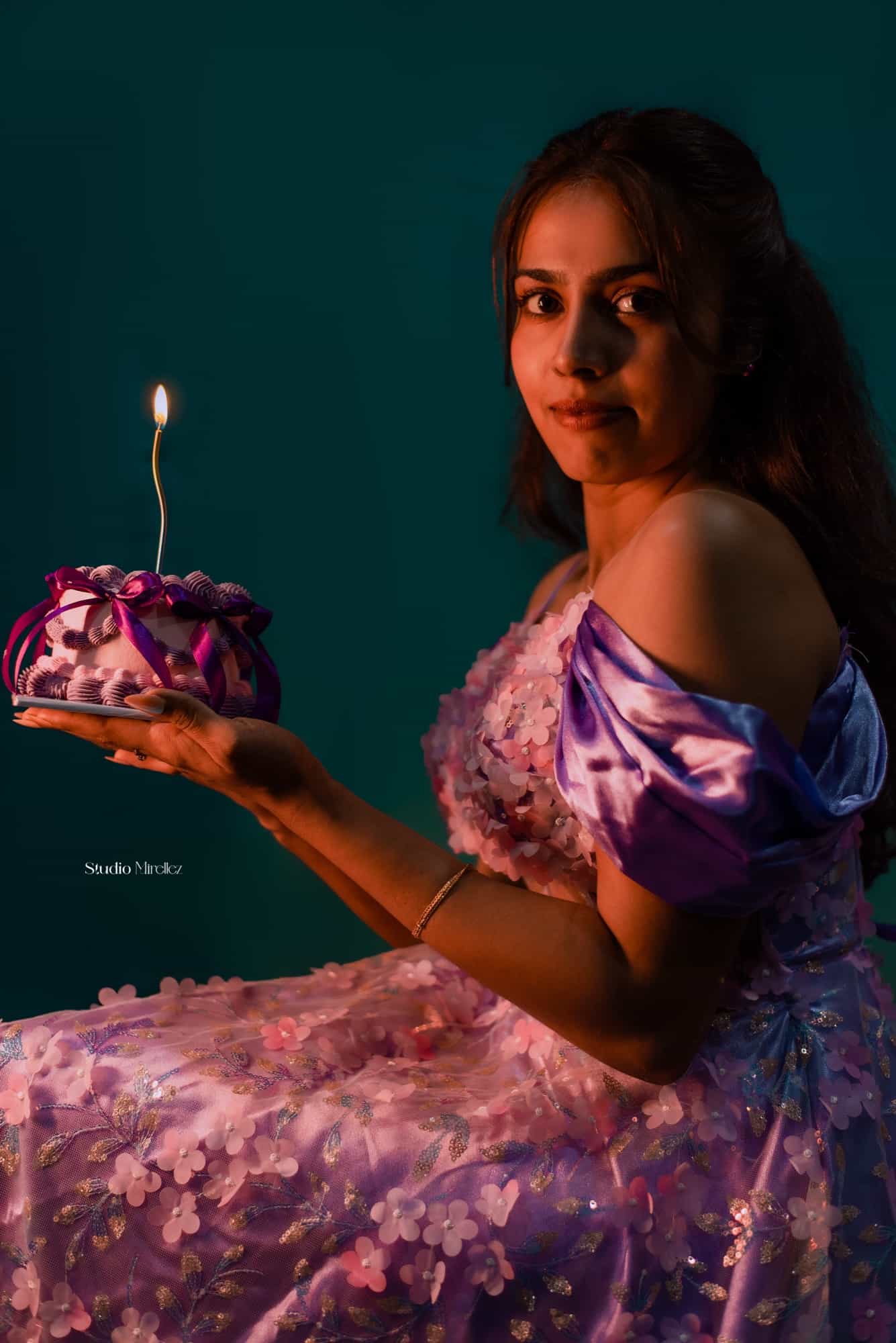 Varangana seated against a deep teal backdrop, holding a ribbon-wrapped birthday cake with a single lit candle