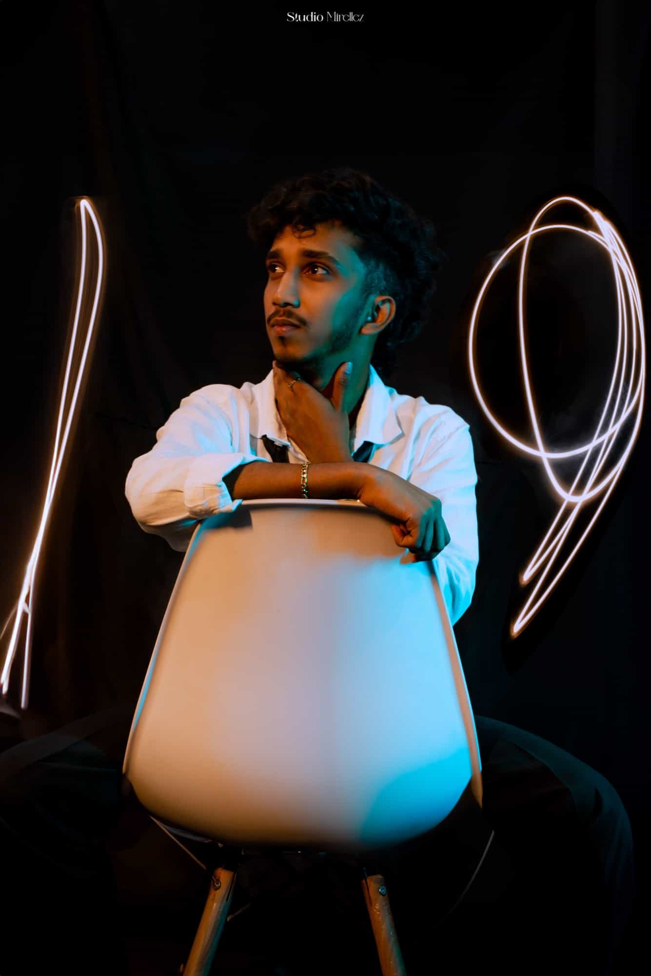 Vihanga sits backwards on a white chair against black, framed by swirling light-painting trails in amber and teal