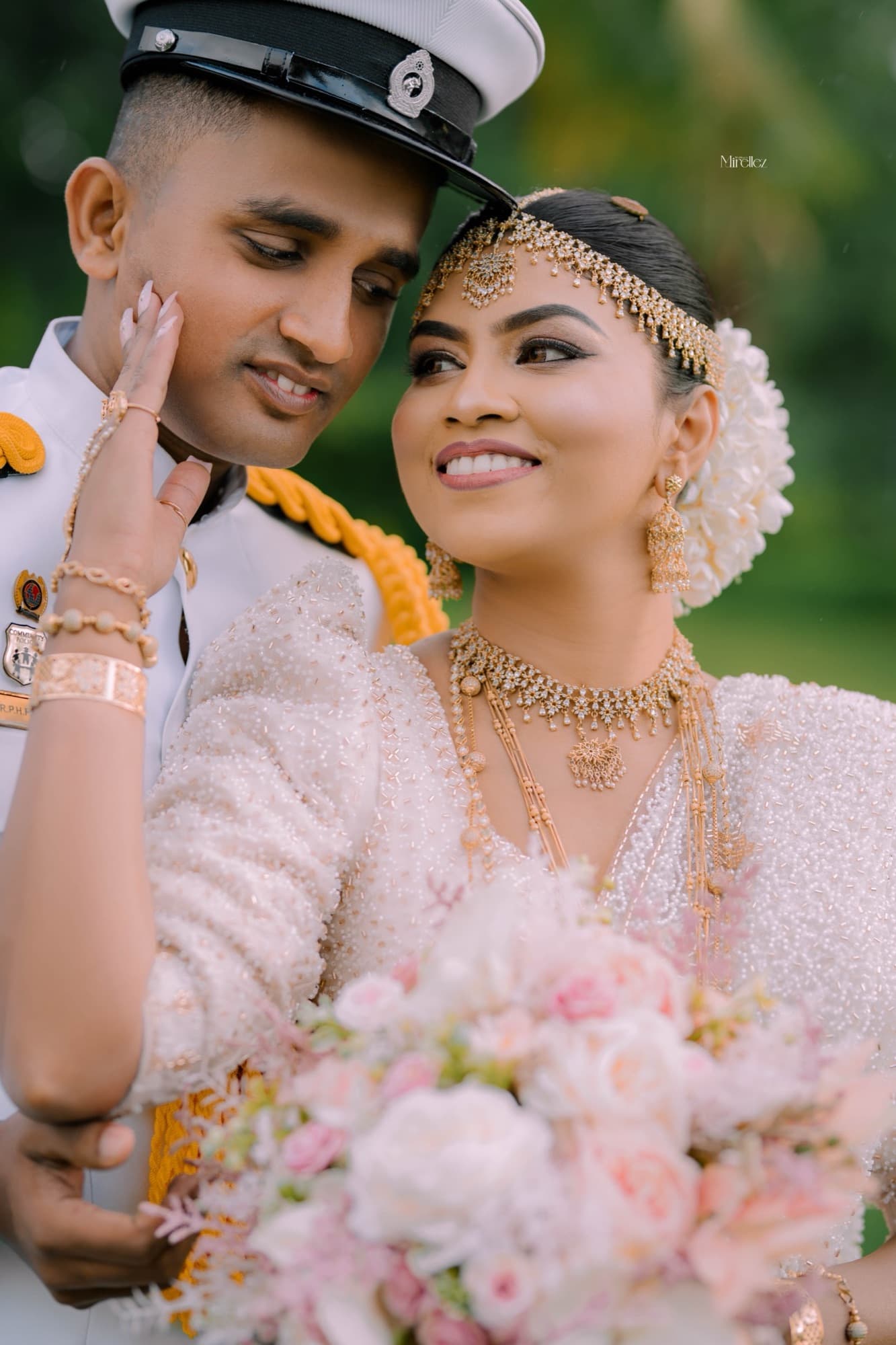 Close-up of the bride tenderly touching the groom's face, her gold Kandyan headpiece and jewellery catching the light as she holds a soft pink bouquet.