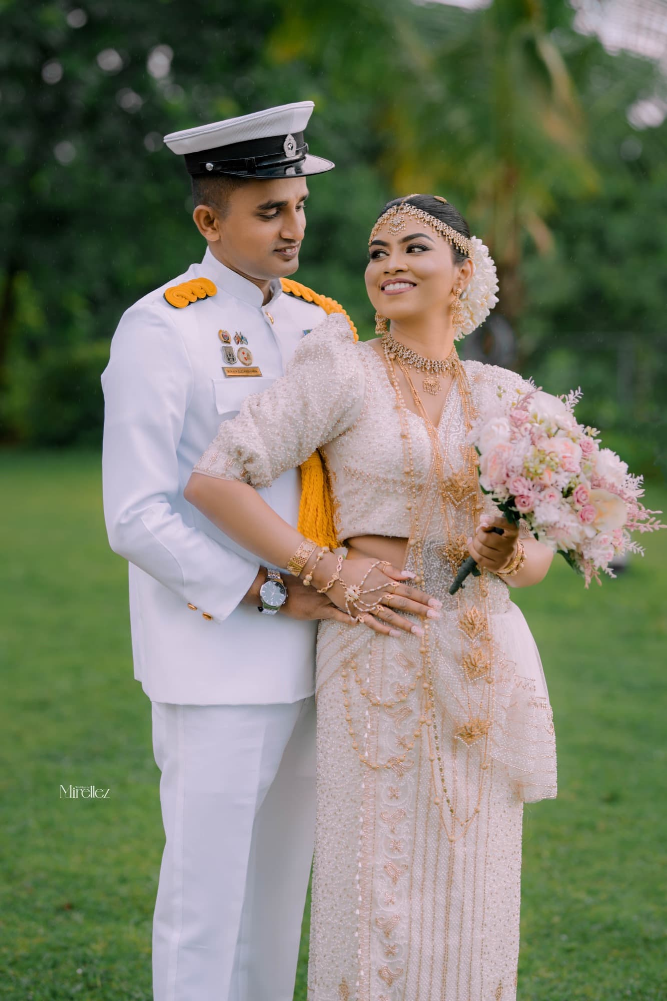 Bride in a pearl-beaded Kandyan saree leans back against her groom in white naval dress uniform, rain falling across a green lawn