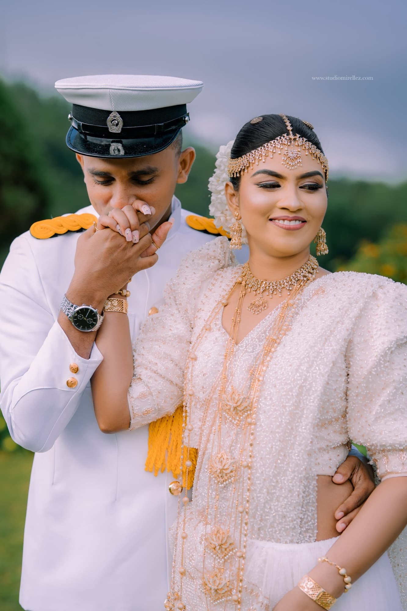 The groom in his white naval dress uniform kisses the bride's hand, his cap tilted low, with her gold-embroidered bridal dress glowing in soft daylight.