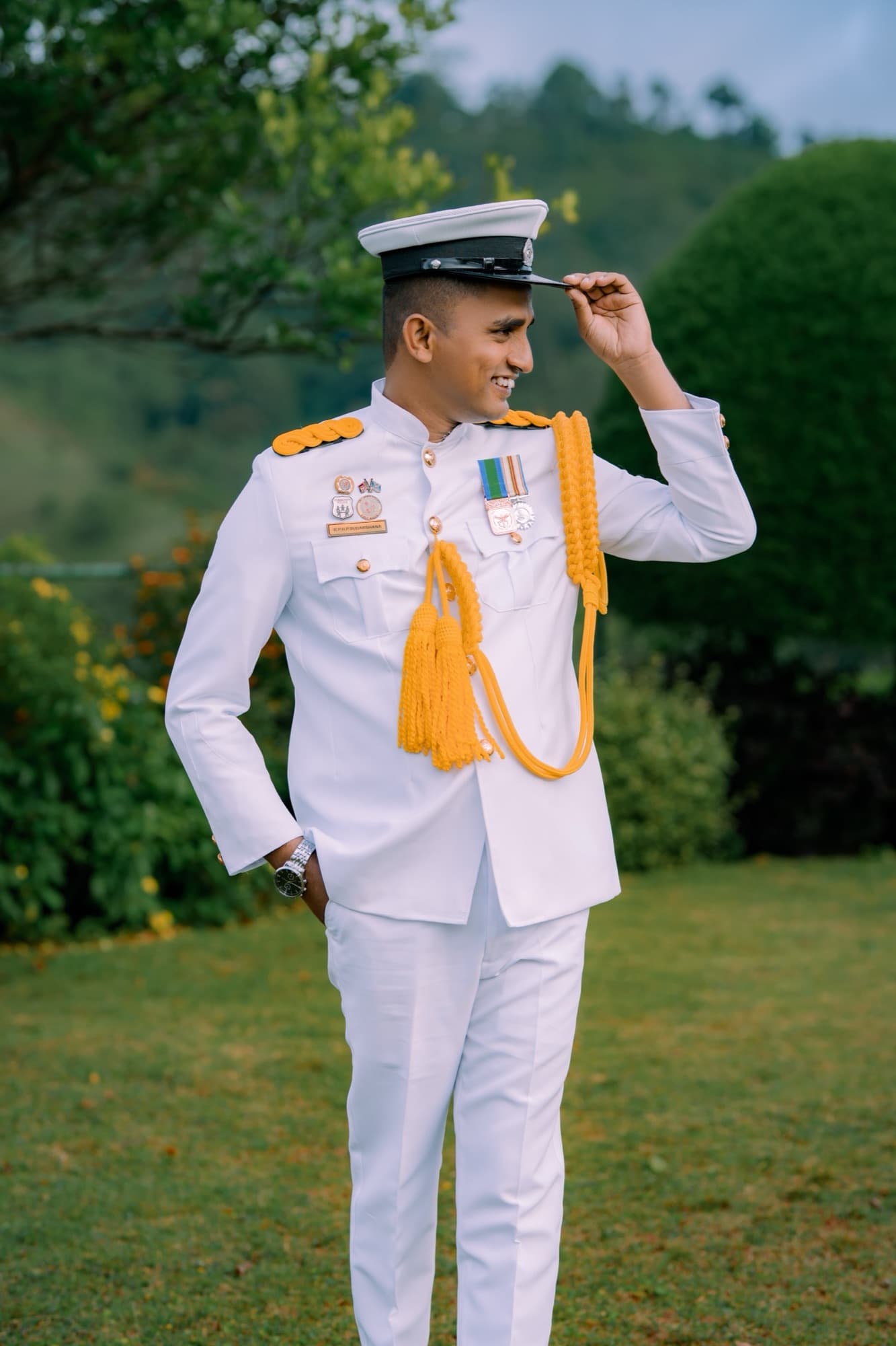 Full-length portrait of the groom in white naval uniform with gold aiguillette and medals, tipping his peaked cap against a lush green garden.