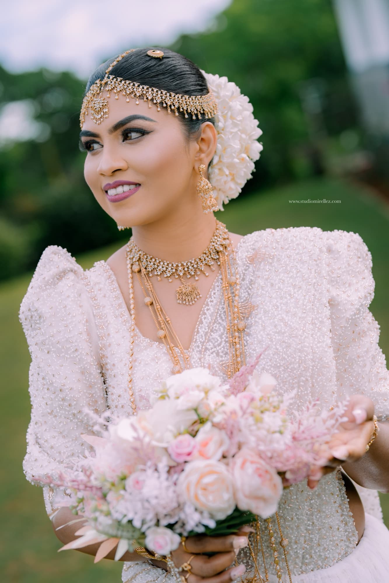 Radiant bridal portrait of the bride in a beaded Kandyan dress and layered gold jewellery, smiling as she cradles a blush pink and cream bouquet.