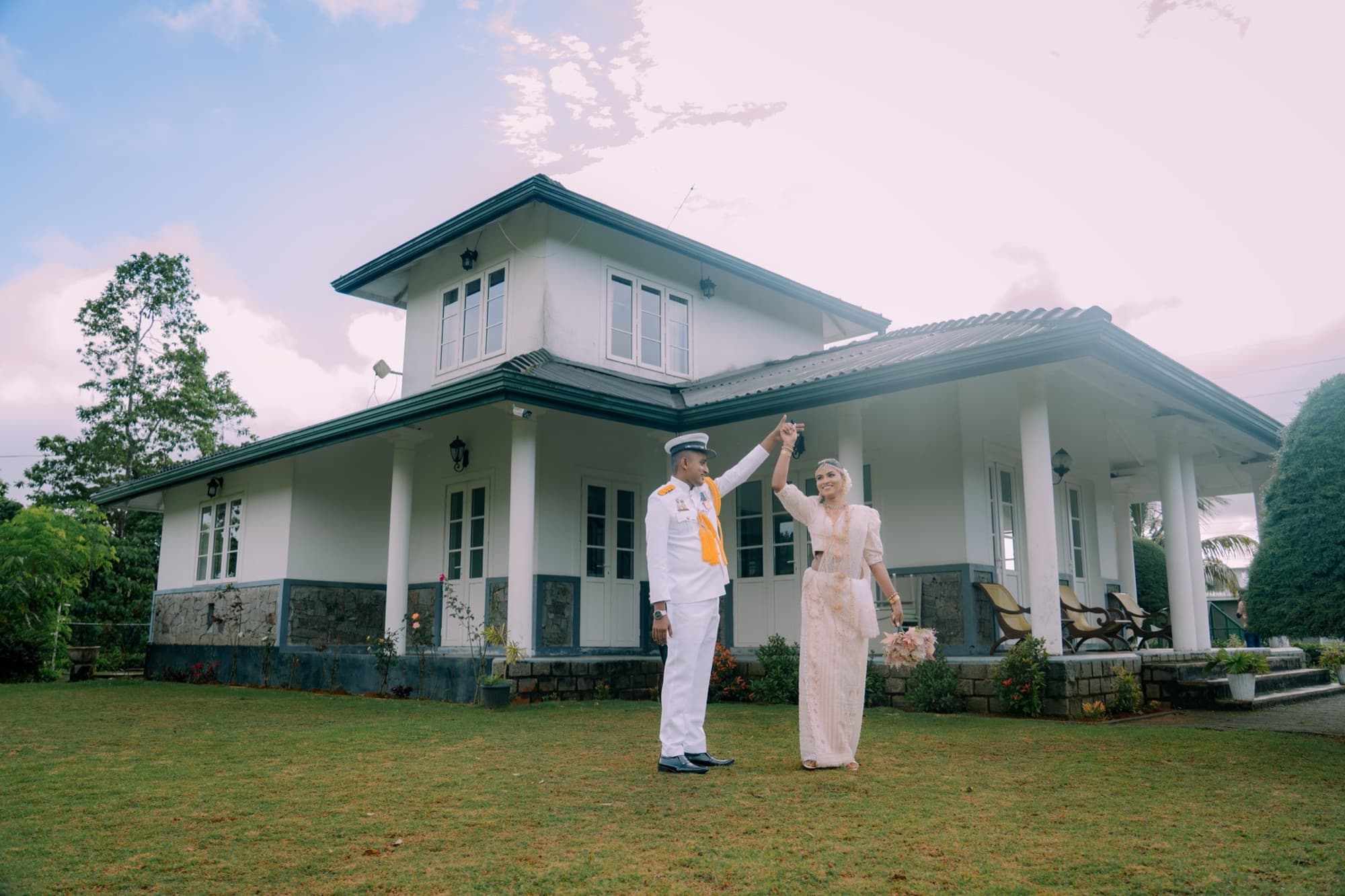 Wide environmental frame of the couple dancing on the lawn in front of a white colonial-style villa under a soft evening sky.