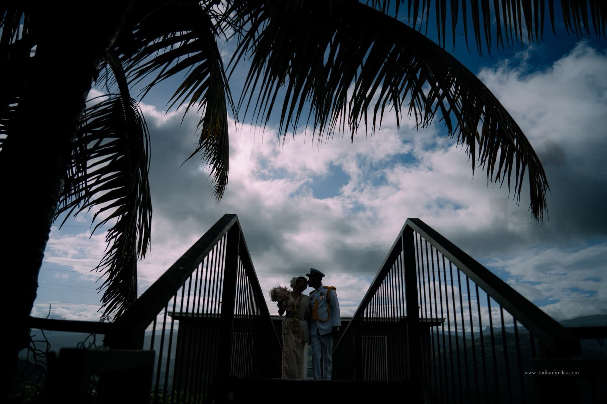 Dramatic silhouette of the bride and groom framed by a coconut palm frond and gabled rooftops against a brooding cloud-filled sky.