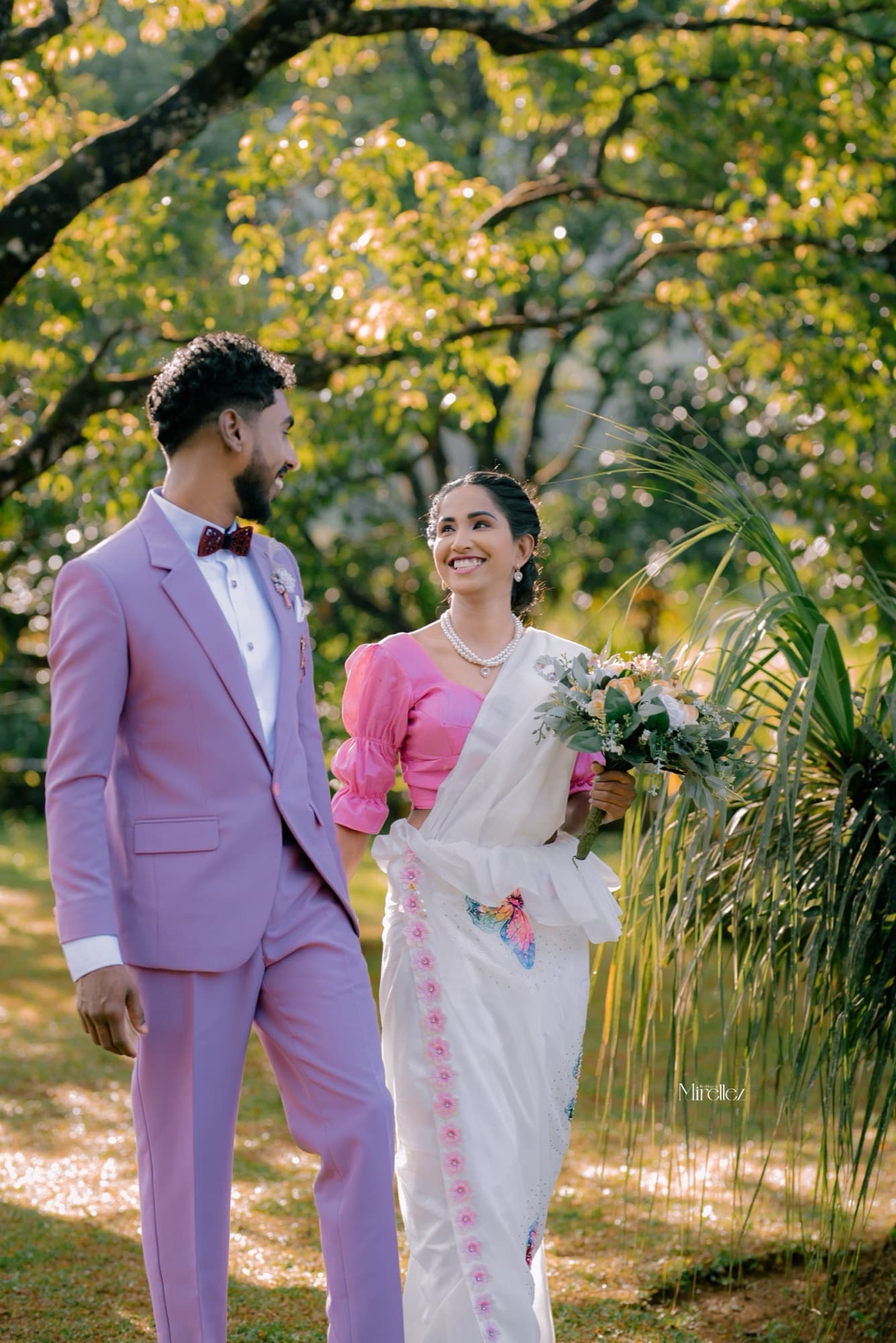 Bride in a pink-and-white saree laughing with her groom against a lush green canopy of morning light
