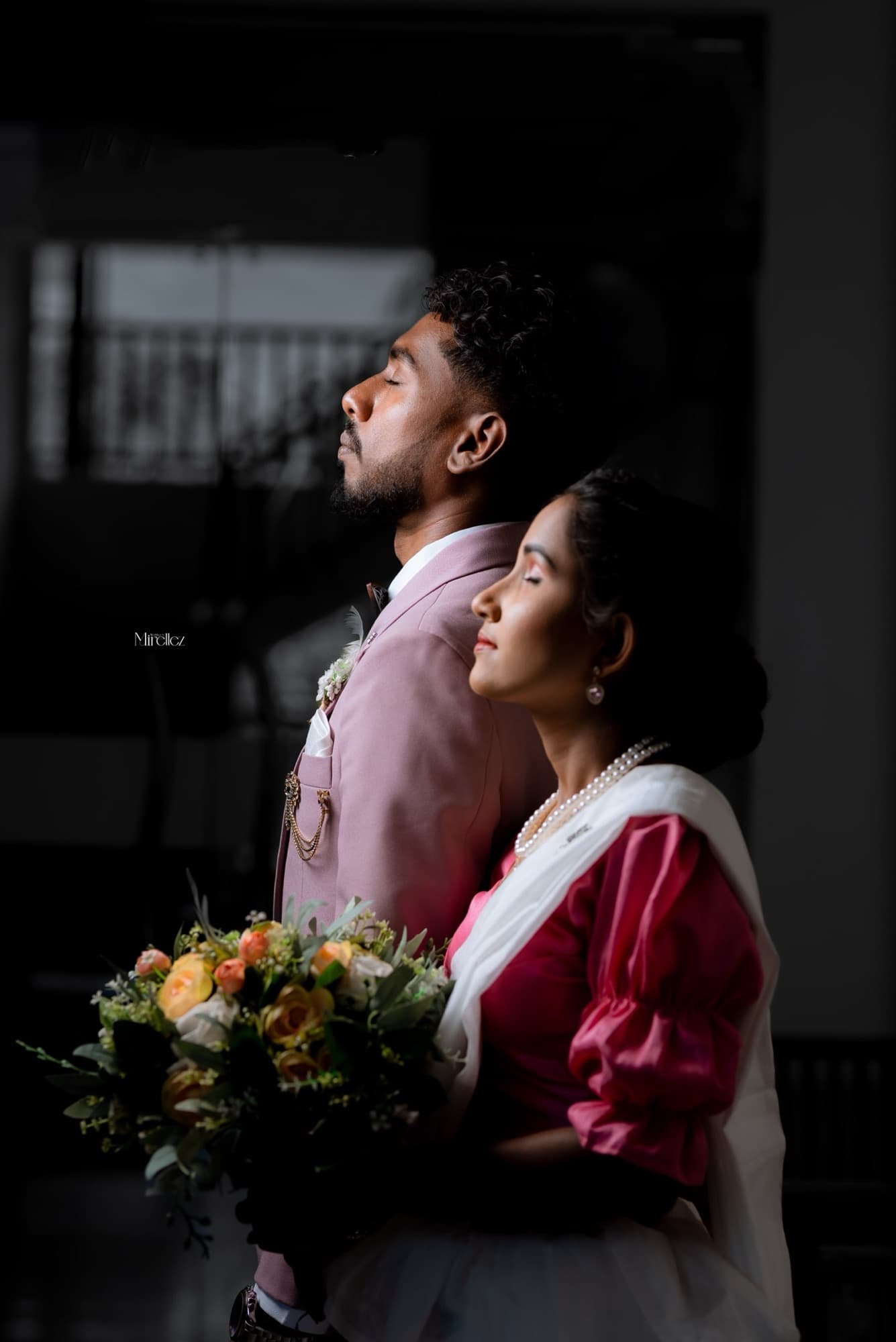Dramatic backlit profile of the couple standing close, the bride's bouquet glowing in low window light