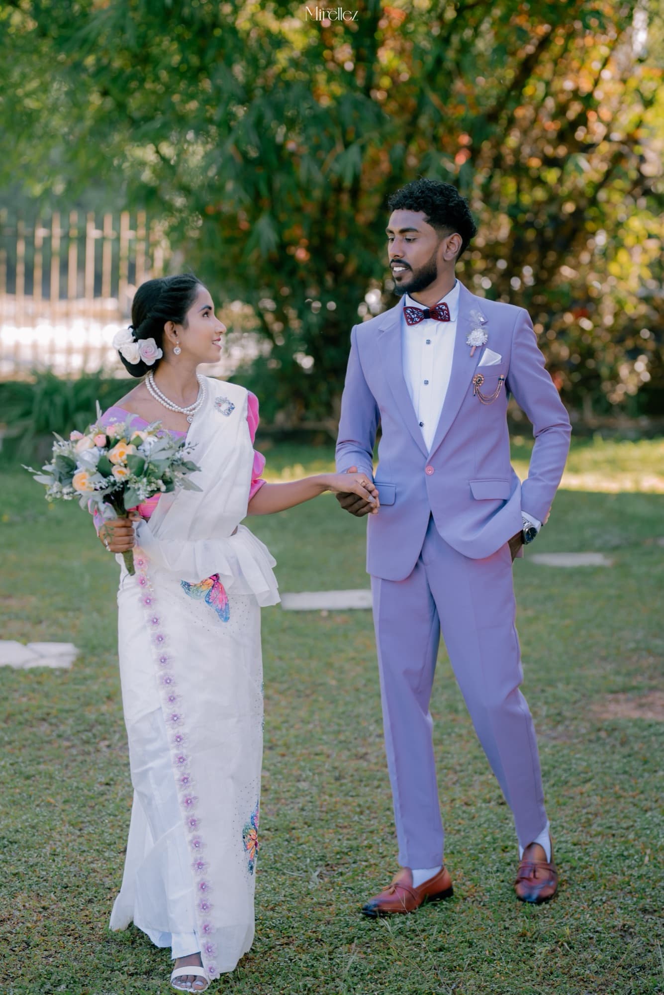 Couple holding hands in a garden, the bride with a flower in her hair and the groom in a blue suit