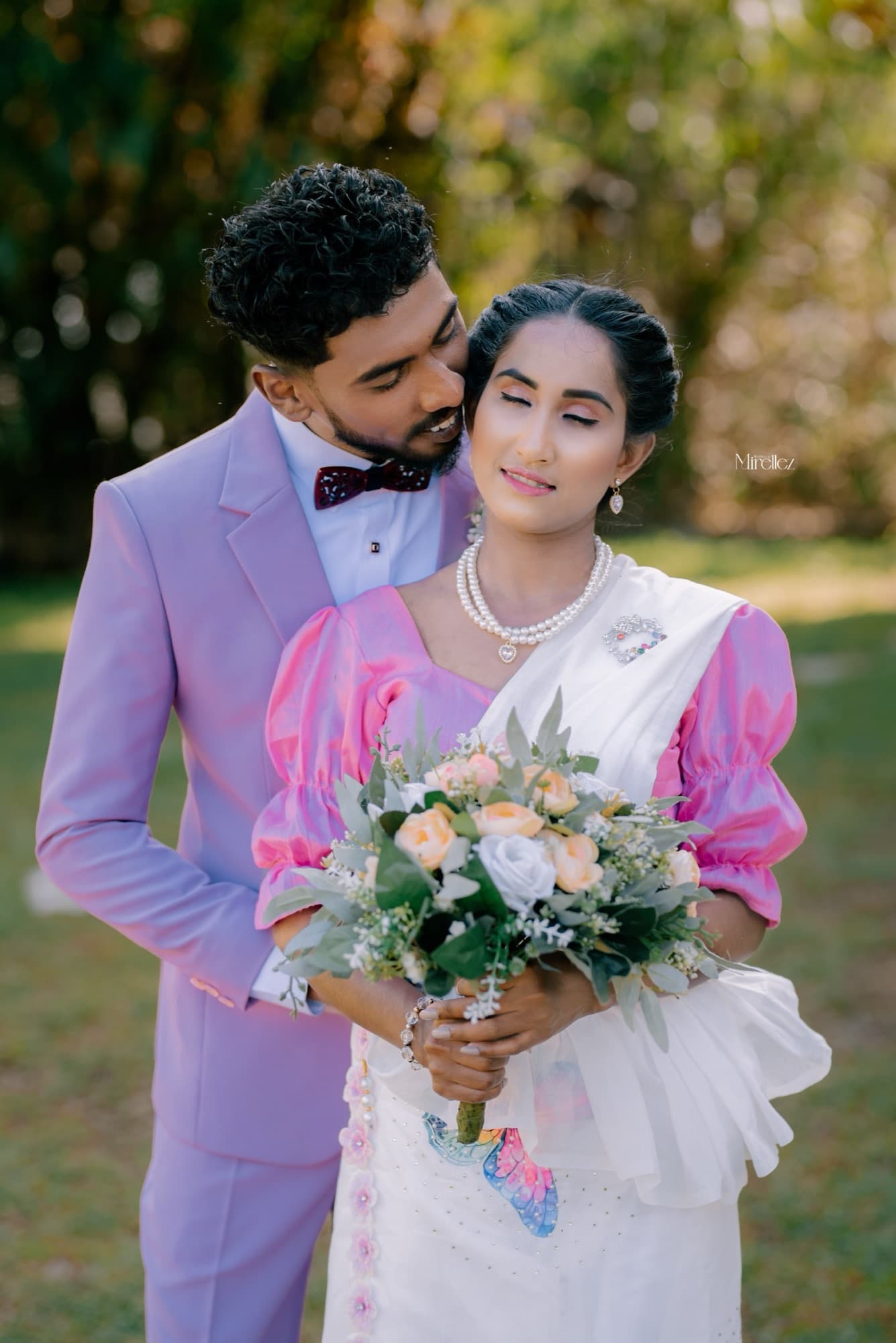 Groom in a lilac suit tenderly kissing his bride's forehead as she holds a soft pastel bouquet in a sunlit garden