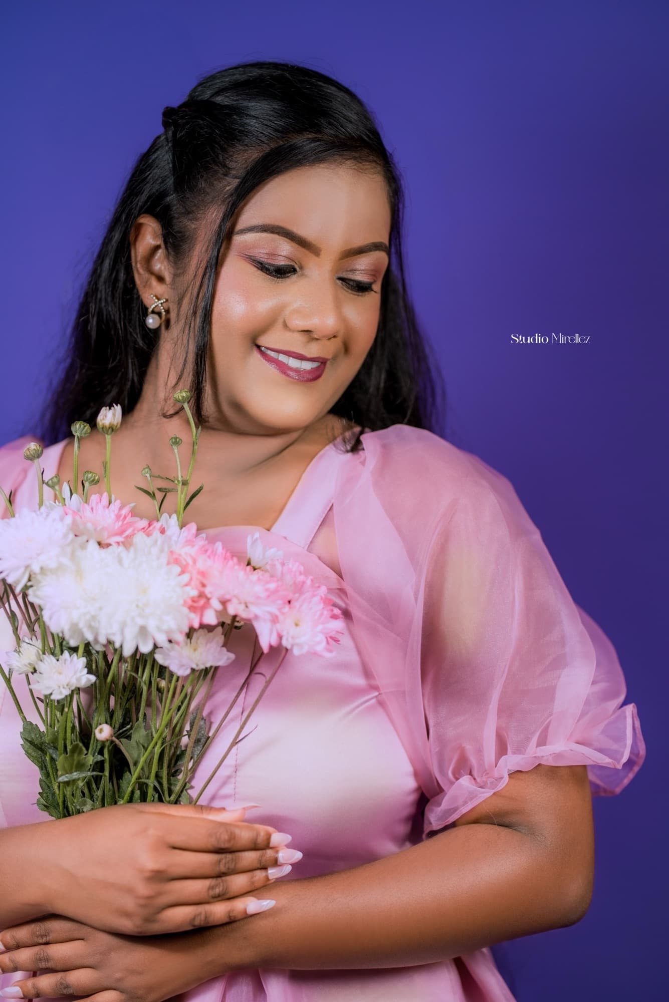 Close-up of a smiling woman in soft pink looking down at a pastel bouquet of pink and white chrysanthemums, gentle studio light