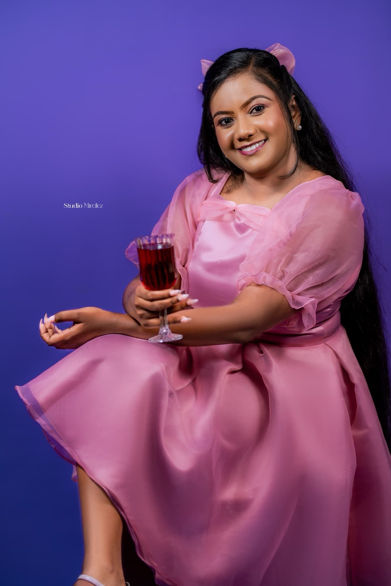 Woman in a pink organza dress seated and smiling to camera, holding a glass of red drink, against a violet studio backdrop