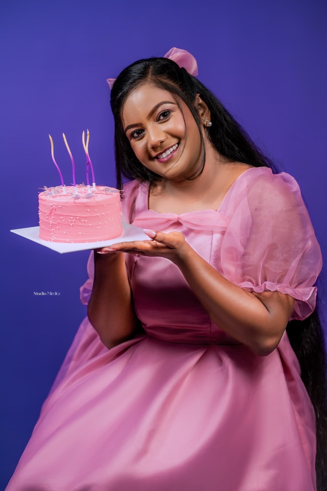 Woman in pink presenting a small pink birthday cake topped with unlit candles, smiling brightly at the camera