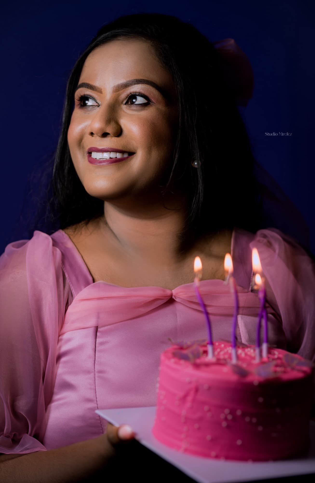 Dramatic low-key close-up of a smiling woman beside a pink cake with glowing lit candles, deep blue background