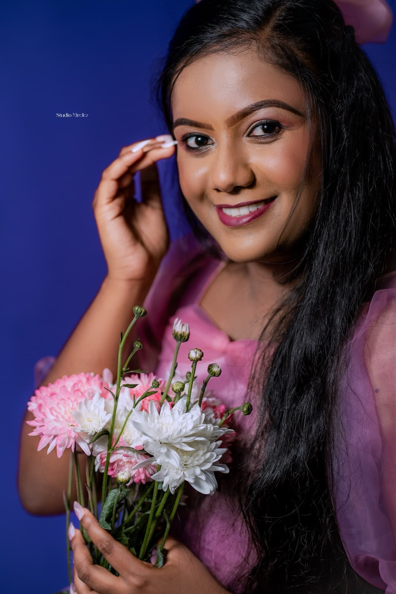 Warm close-up portrait of a woman in pink resting a hand near her face above a pastel bouquet, soft smile to camera