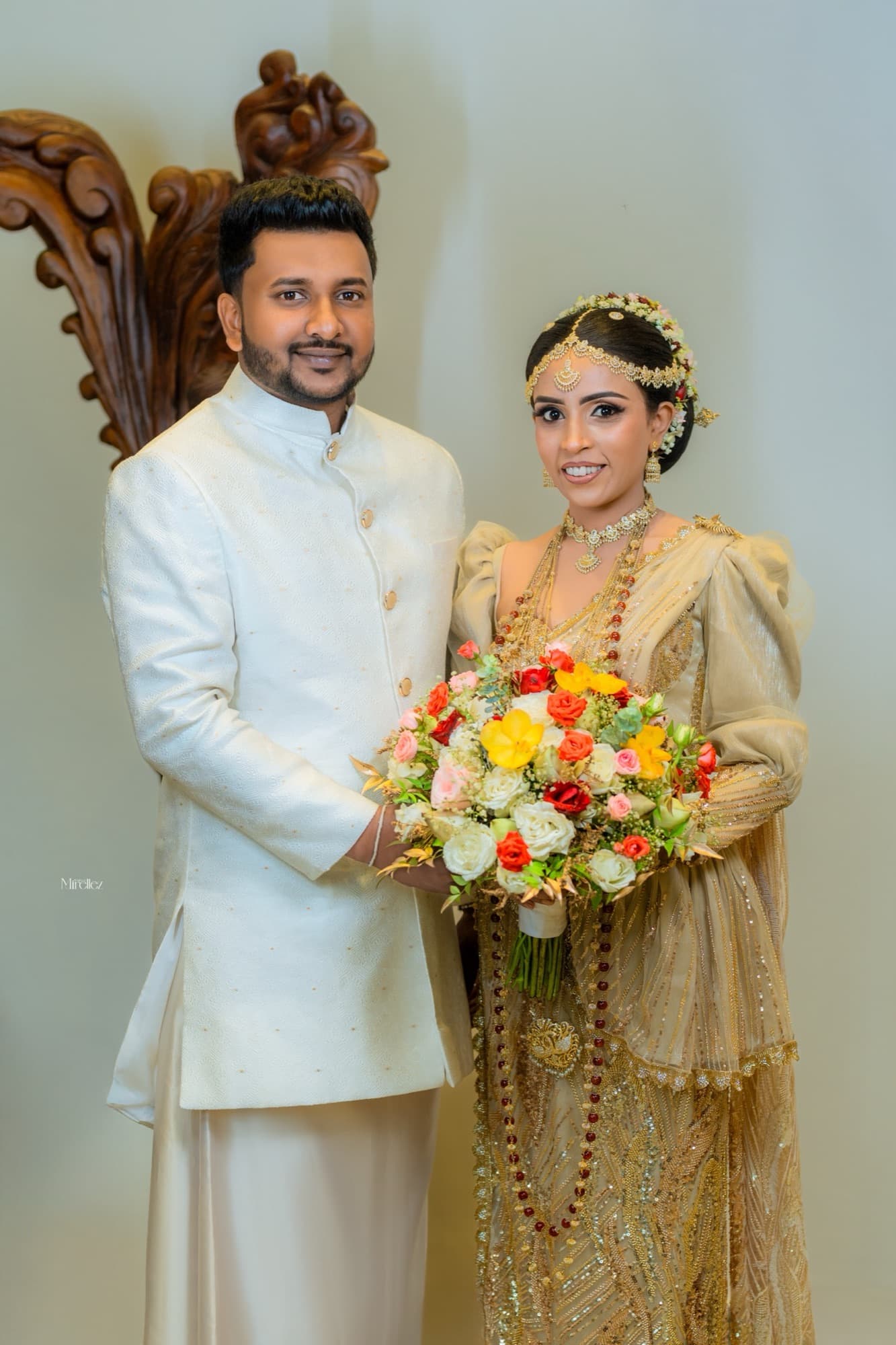 Formal full-length portrait of the couple in traditional Sri Lankan wedding dress holding the bridal bouquet before a carved wooden backdrop.
