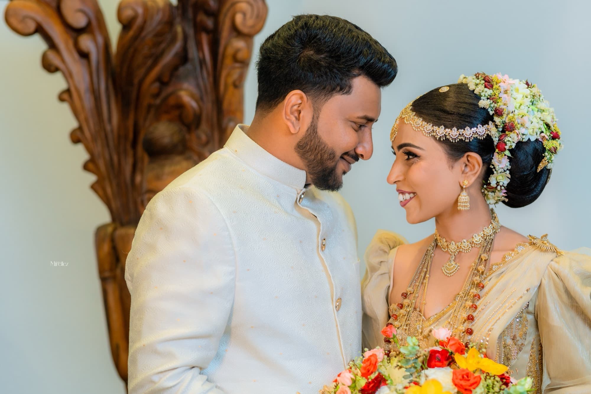 Bride and groom in traditional Sri Lankan wedding attire gazing at each other in soft natural light, the bride in a gold saree with a floral headpiece.