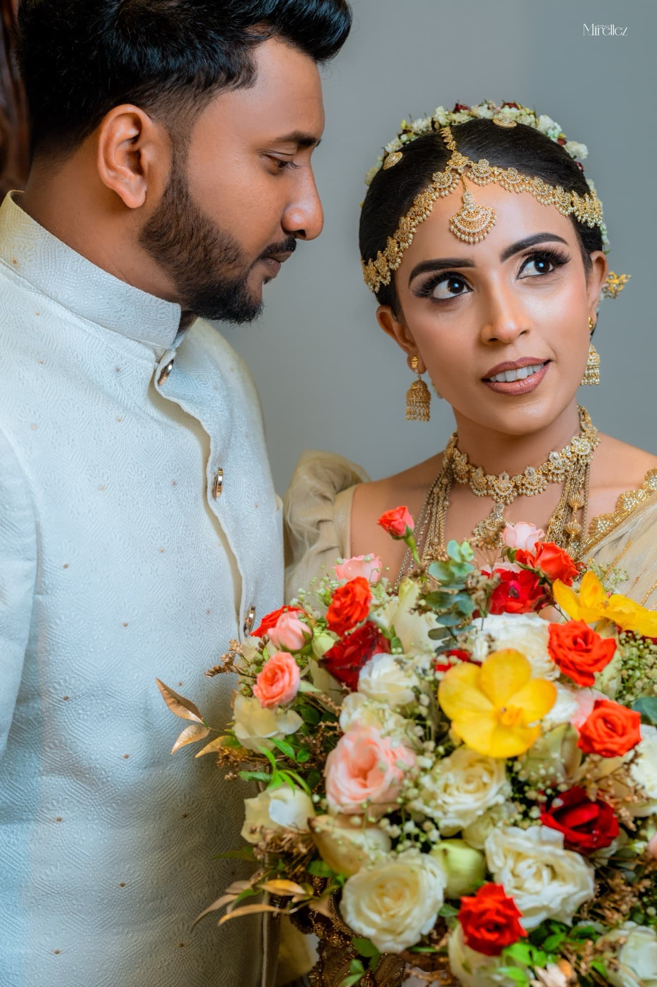 Intimate portrait of the bride looking to camera over a vivid red, yellow and white bouquet, her gold jewellery and headpiece catching the light beside the groom.