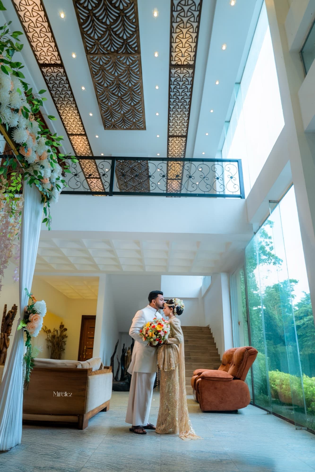 Wide architectural shot of the couple embracing in a grand sunlit villa lobby beneath an ornate patterned ceiling and floral arch.