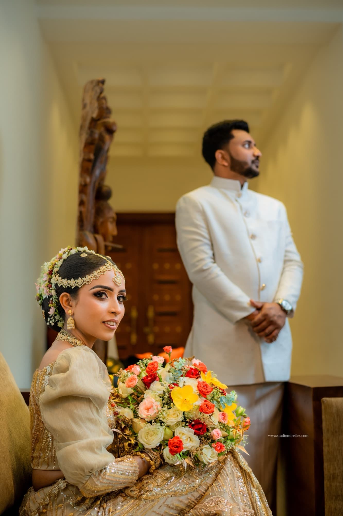 Editorial frame of the seated bride holding her bouquet and glancing back, with the groom standing softly out of focus behind her.