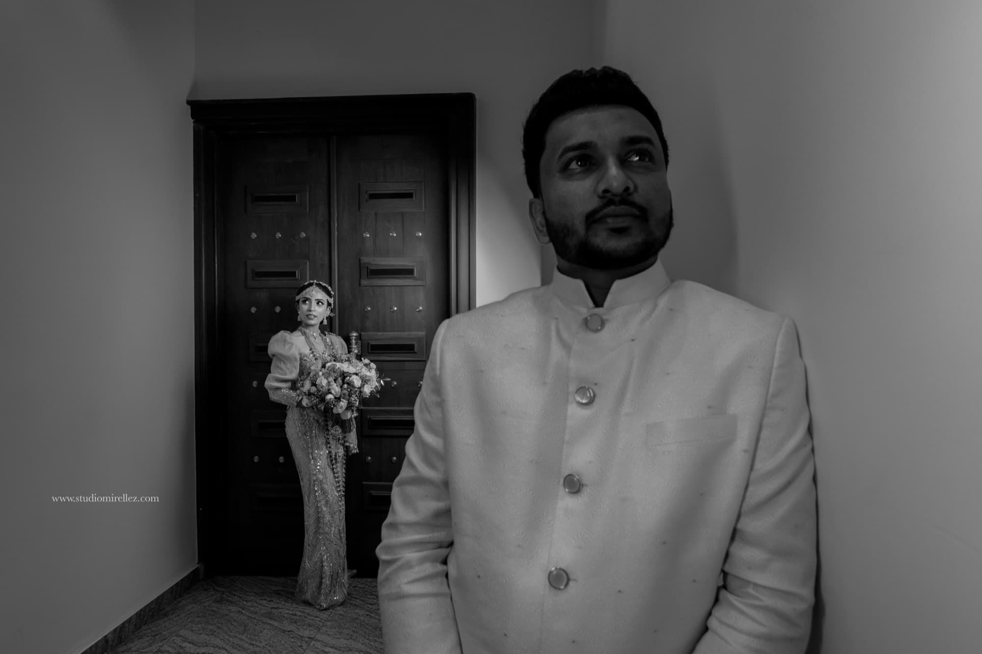 Cinematic storytelling frame with the groom in the warm-lit foreground and the bride framed in a doorway holding her bouquet.
