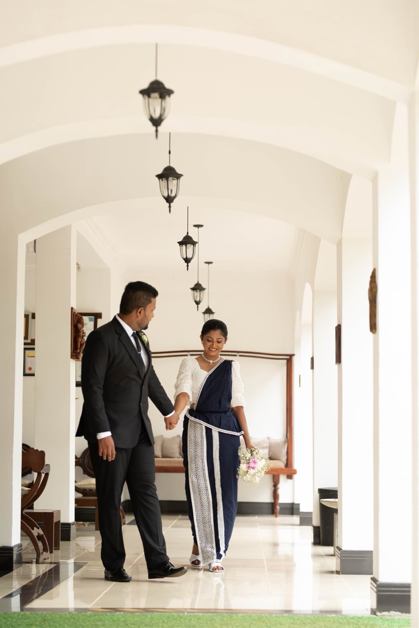 Couple walking hand in hand down a bright white arched colonial hallway lined with hanging lanterns