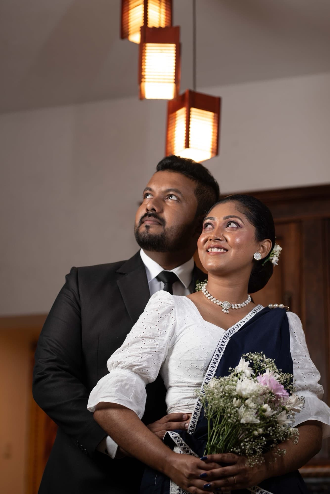 Engaged couple in a close embrace beneath warm glowing lanterns, the bride smiling against the groom's shoulder, holding a soft pastel bouquet