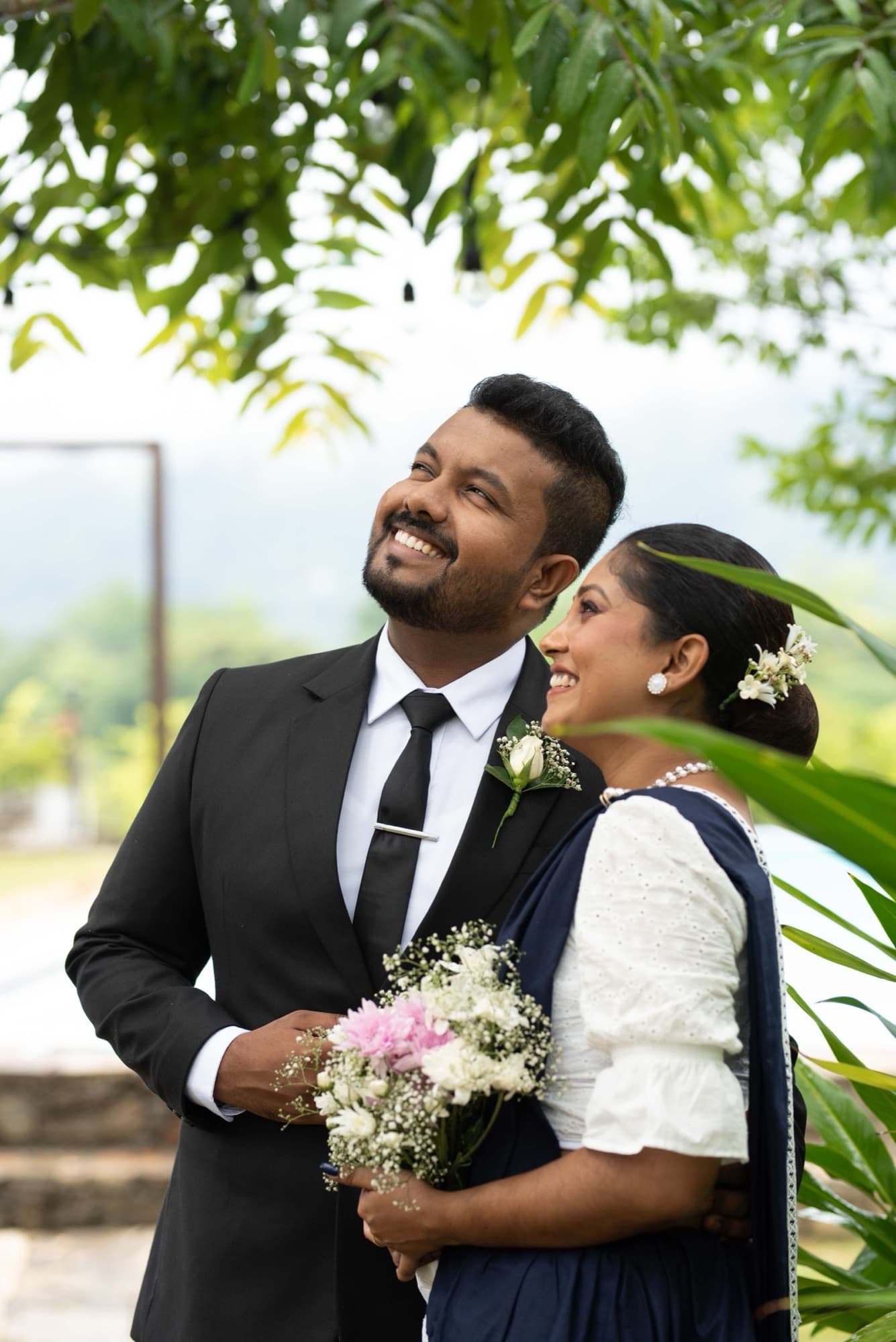 Couple laughing together in a lush green garden, the bride resting against the groom as they hold a white-and-pink bouquet